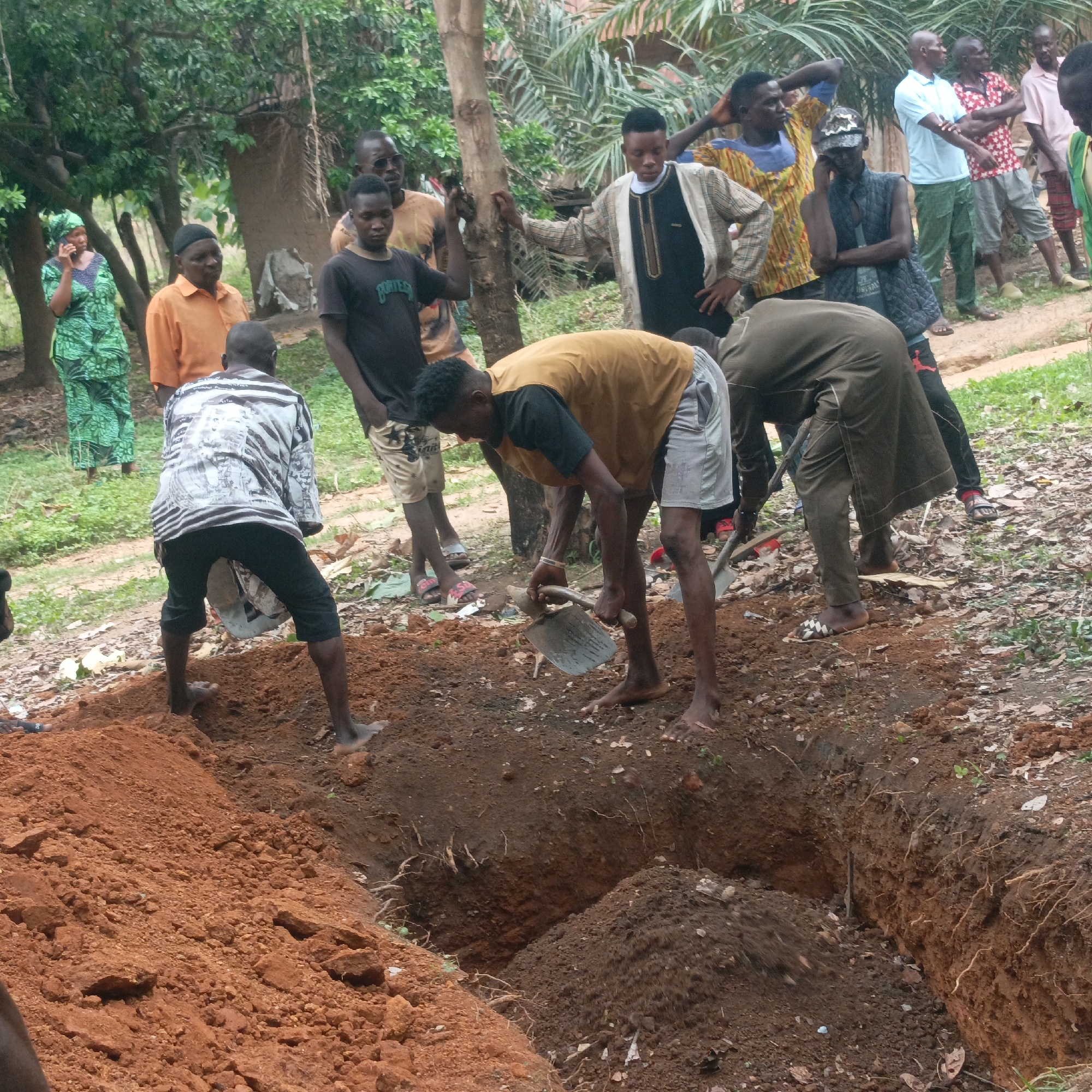 The image depicts a group of people gathered around a freshly dug grave in Andaha, Nigeria. Three individuals are actively digging, using shovels to move soil. The grave is a rectangular pit in reddish-brown earth. A mound of excavated dirt sits beside it. The diggers are barefoot, wearing casual clothing.
Around the grave, other people stand, watching or waiting. They are a mix of men and women, dressed in a variety of clothing, from patterned shirts and dresses to more formal attire. Some are barefoot, others wear sandals. The background shows a rural setting with trees and simple structures.