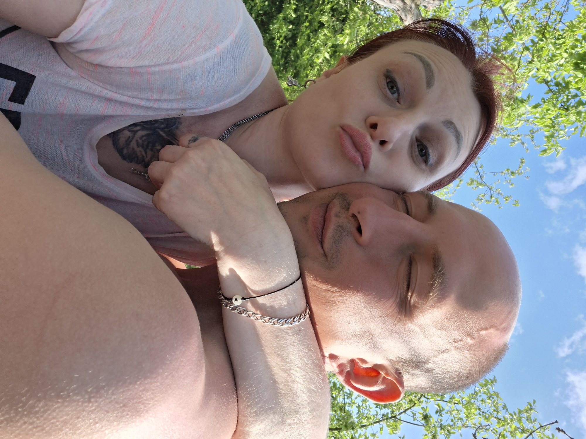 The image shows a man and a woman lying in an outdoor setting, likely a park or yard, in Сваткаўскі сельскі Савет, Belarus. The woman, with auburn hair pulled back, a fair complexion, and distinct eyebrows, is in the foreground with her arm draped around the man's neck. She is wearing a light-colored shirt and a silver chain. She is pouting her lips toward the camera. The man has a shaved head and closed eyes. He is bare-chested. Both are facing upwards toward the sky visible through tree branches. The woman has a tattoo on her chest. The man has a bracelet on his wrist.
