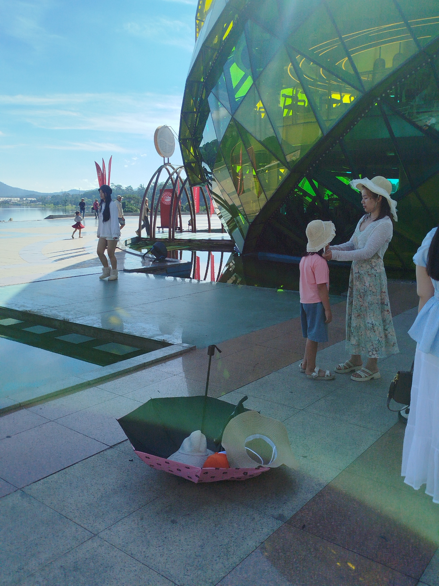The image depicts a sunny day at the Da Lat Flower Garden, Vietnam, specifically near the "Big C" or Lam Vien Square. In the foreground, a partially opened green umbrella lies on the tiled ground, adorned with a pink fabric insert featuring small black heart patterns. Resting inside the umbrella are two straw hats, one a wide-brimmed floppy style and the other a smaller, more circular one. To the right, a woman wearing a white long-sleeved top and a floral print skirt, along with a white straw hat, is adjusting the hat of a child in a pink t-shirt and blue shorts, who is also wearing a straw hat. Behind them, a distinctive geodesic dome-shaped building with green, faceted glass panels dominates the background, reflecting the sky and surrounding environment. Further in the distance, several individuals are visible walking along a paved area next to a body of water, with a hilly landscape and a bright blue sky with wispy clouds overhead. Red banners with an emblem are also present, attached to metal structures. The overall scene suggests a recreational area or tourist attraction in Da Lat.