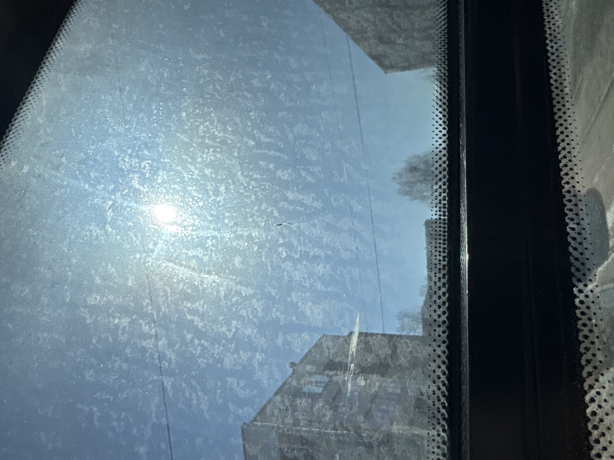 The image shows a view through a window with a frosty, speckled texture obscuring the scene. The sun's glare is visible through the frost, slightly off-center to the left. Outside the window, blurred shapes suggest buildings, including one with a pitched roof and another with a flat roof, consistent with the urban architecture found in Berezniki, Russia. The window frame is black, with a dotted edge. There is no discernible human presence.