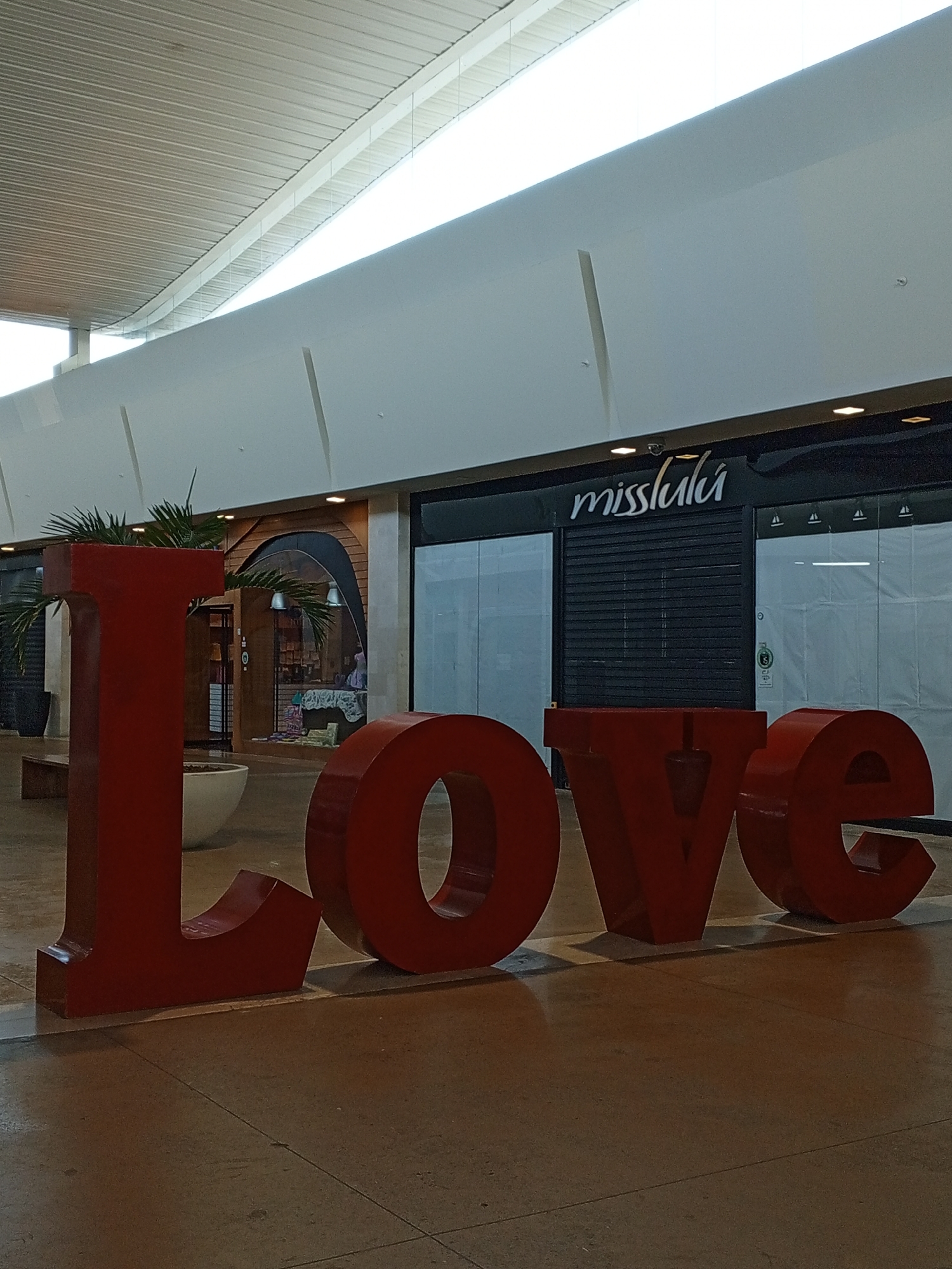 The image shows a large, red "LOVE" sculpture in front of storefronts in what appears to be a shopping mall. One of the stores is named "misslula" and has its security gate pulled down. There's another store with a wooden framed doorway to the left. The location is Pampatar, Venezuela, based on additional context (not visible in the image). The indoor environment has a high, modern ceiling with linear lighting. The floor is smooth and appears to be made of polished concrete. There are no people clearly visible in the frame.