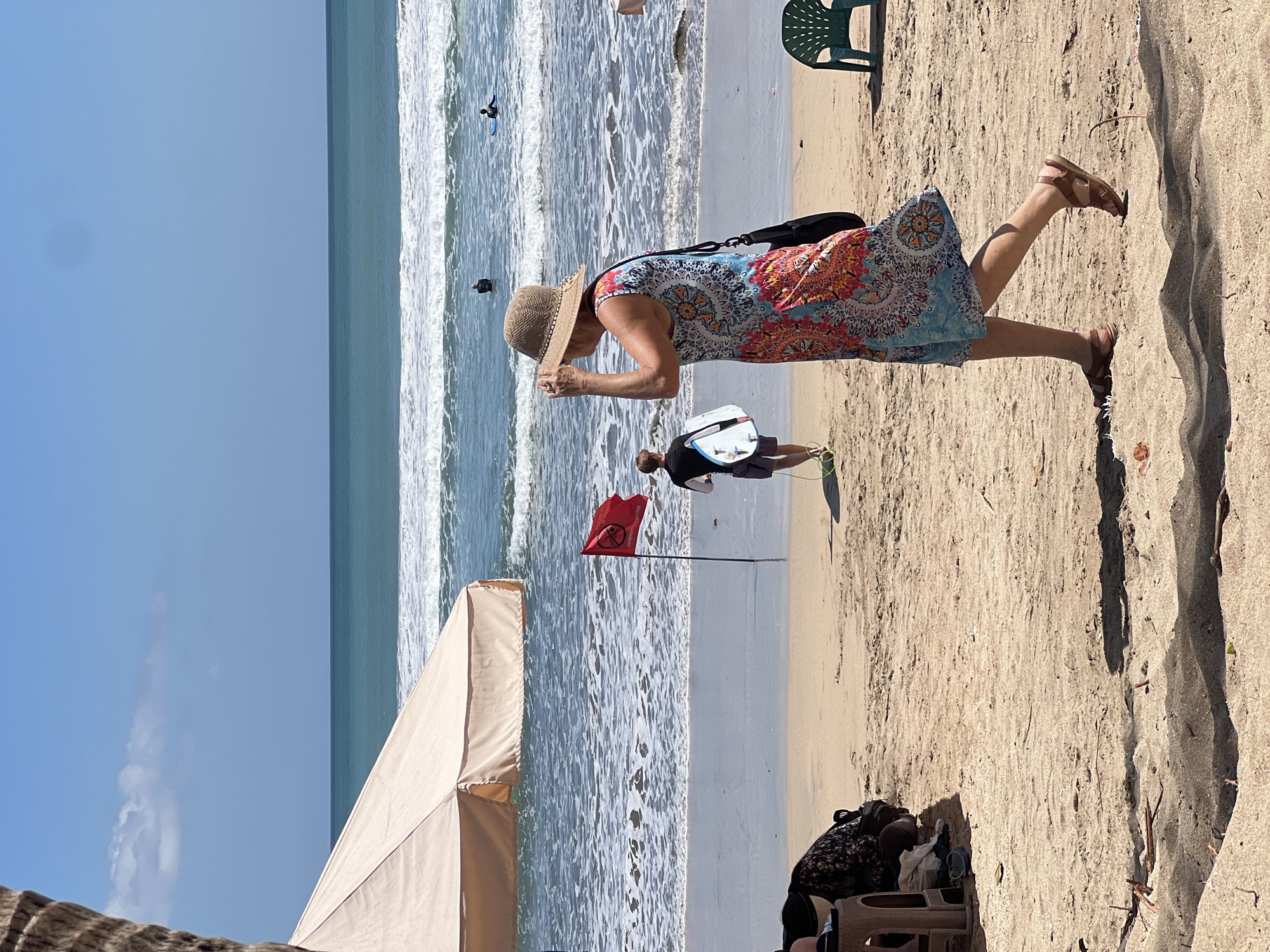 On a sunny day at Kuta Beach, Indonesia, a woman wearing a colorful floral dress and a straw hat walks along the sandy shore. She is facing away from the camera, her right hand adjusting her hat. To her left, a young person in a black rash guard and shorts walks towards the ocean carrying a surfboard. In the background, the turquoise ocean has moderate waves, with several surfers scattered in the water. A red flag with a warning symbol and text indicating "SWIMMING PROHIBITED" is planted in the sand near the waterline. To the left of the frame, part of a large, beige beach umbrella is visible, providing shade. Scattered on the sand are some personal belongings, including a patterned bag. Further to the right, a green plastic beach chair sits unoccupied. The overall scene depicts a lively beach environment with clear skies and a gentle breeze.