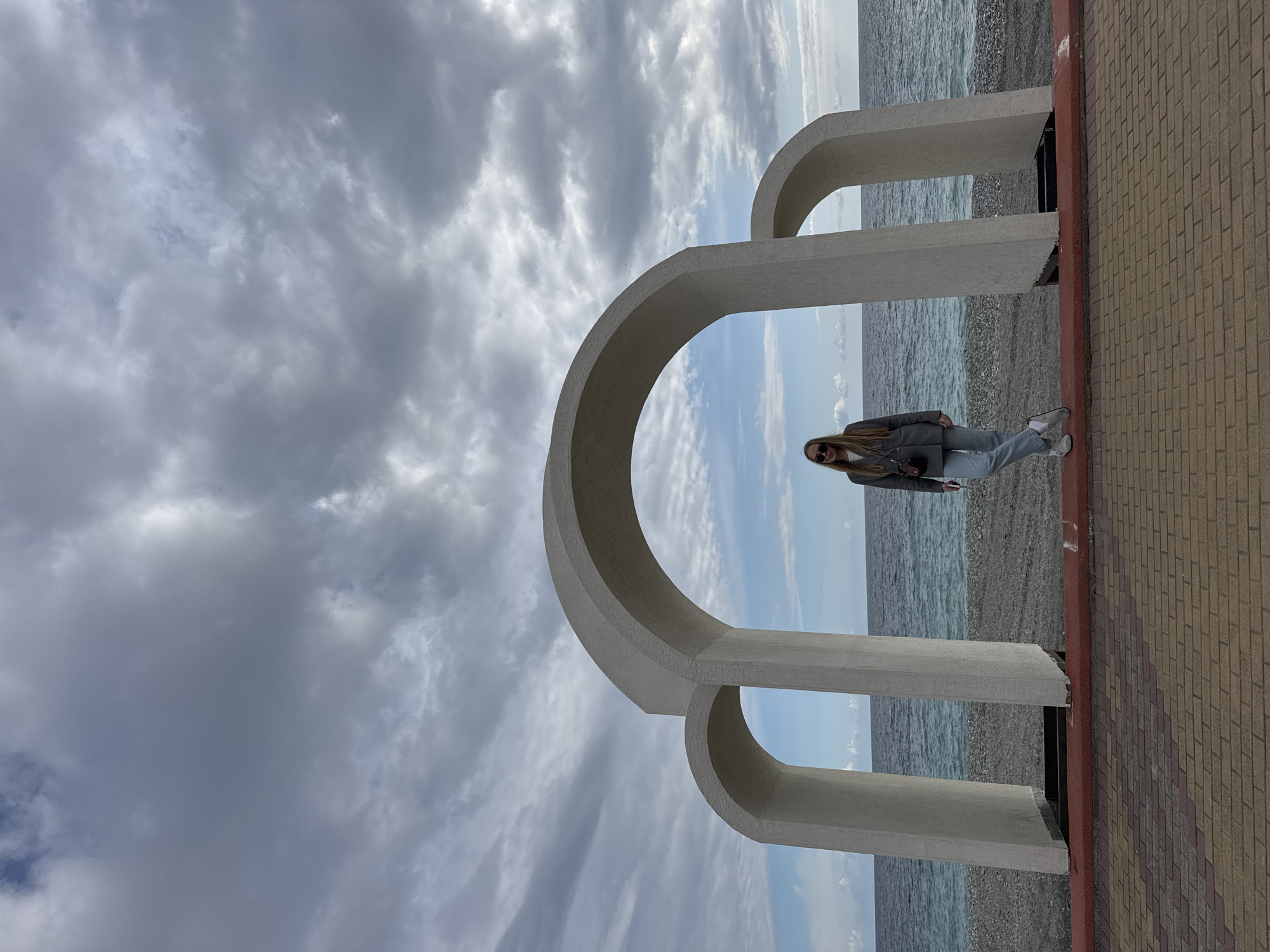 The image depicts a woman standing near a large architectural structure in Sirius, Russia. The structure consists of three interconnected, rounded arches made of a light-colored material, possibly concrete. The arches frame a view of the sea, visible through the gaps. The woman is wearing a gray blazer, light-colored jeans, and sunglasses. She is standing on a brick pathway that runs alongside the structure. The background shows a cloudy sky above the sea. The ground between the pathway and the sea appears to be pebbled. The photograph appears to be taken during daylight hours.