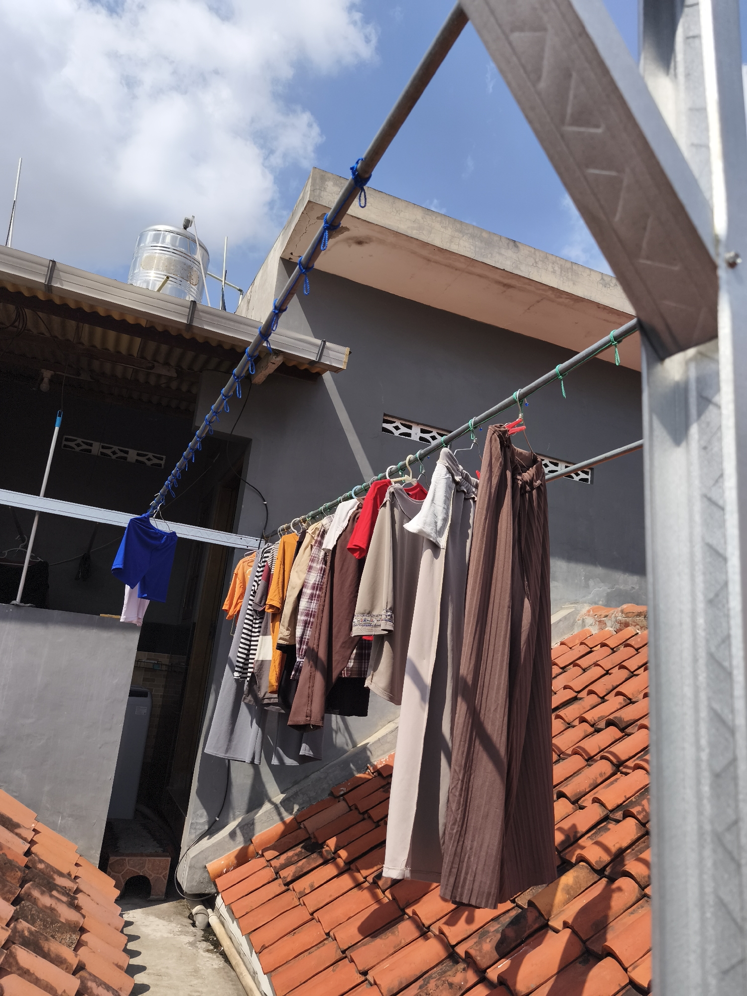 This image captures a rooftop scene in Yogyakarta, Indonesia, where laundry is drying on a clothesline. The foreground is dominated by a section of orange tiled roofing, casting shadows from the hanging garments. In the mid-ground, various pieces of clothing, including shirts, pants, and a long brown skirt, are suspended on a metal clothesline supported by blue and green ropes attached to a building structure. Behind the clothesline, a dark-colored building with a large window or opening is visible. To the left, a silver water tank is situated on the roof, next to a corrugated metal awning. The sky above is bright blue with scattered white clouds. The overall impression is one of everyday domestic activity taking place on a rooftop in a tropical urban environment.