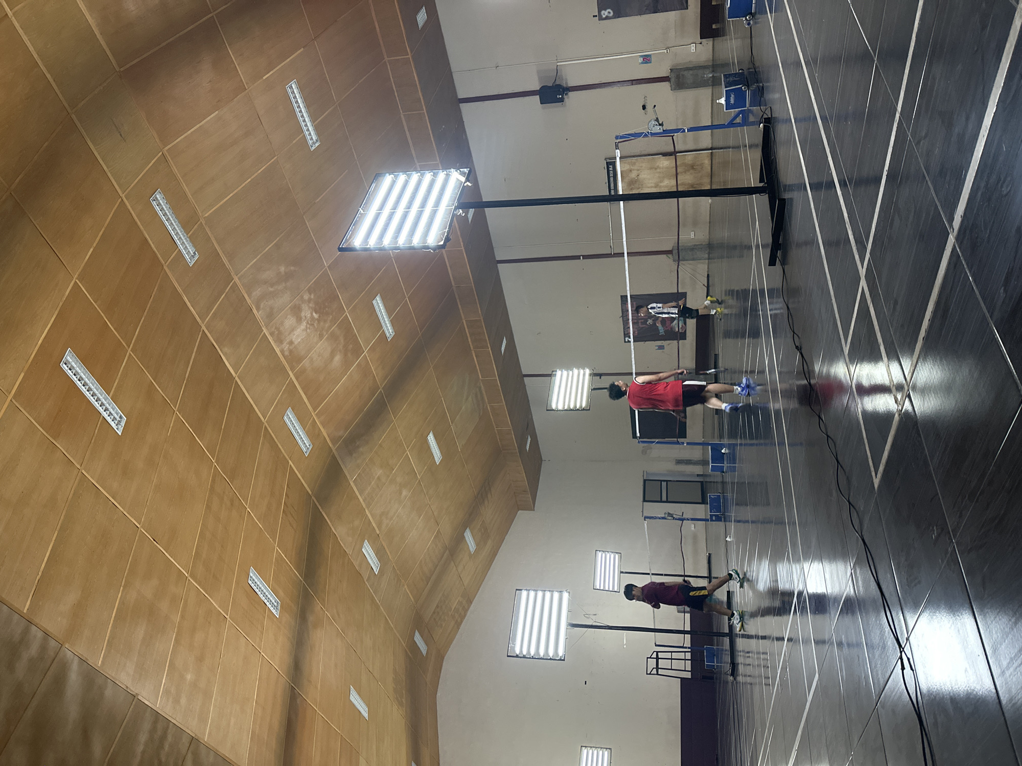 An indoor badminton court in Batu, Indonesia, is depicted, featuring a polished, dark floor marked with white lines. The ceiling is constructed of wooden panels with numerous fluorescent lights. Three individuals are visible on the court. Two players are actively engaged in a game, one in a red shirt and shorts, the other in a black and white striped shirt and shorts. A third person, wearing a maroon shirt and dark shorts, walks across the court. Several portable, bright LED lights are positioned on stands around the court, illuminating the playing area. Badminton nets and posts divide the court, and a portable umpire's chair is visible in the background. The environment appears to be a sports facility.