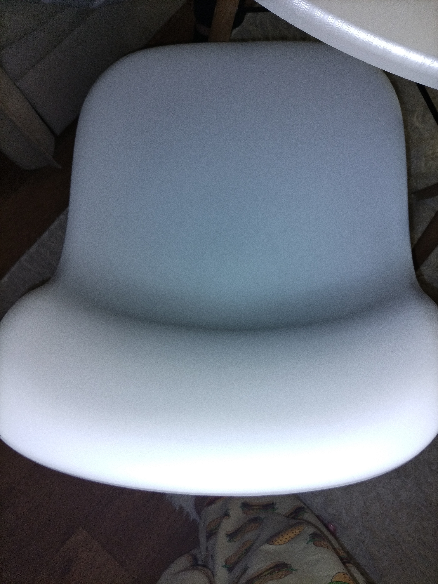 An overhead view of a white molded plastic chair seat is the primary subject of the image. The chair seat has a curved, ergonomic design with a slightly raised back and a wider front. The material appears to be smooth and matte, reflecting the light from above. The chair is positioned on a patterned rug, which is visible at the bottom and sides of the image. In the upper left corner, a portion of another upholstered piece of furniture, possibly a sofa or armchair, is visible. A bright, translucent object, possibly a lamp or a diffuser, is partially visible at the top of the image, casting a strong light. The background is a mix of the rug, the other furniture, and some wooden flooring. The scene is indoors, with artificial lighting. There are no people or animals present in the image.