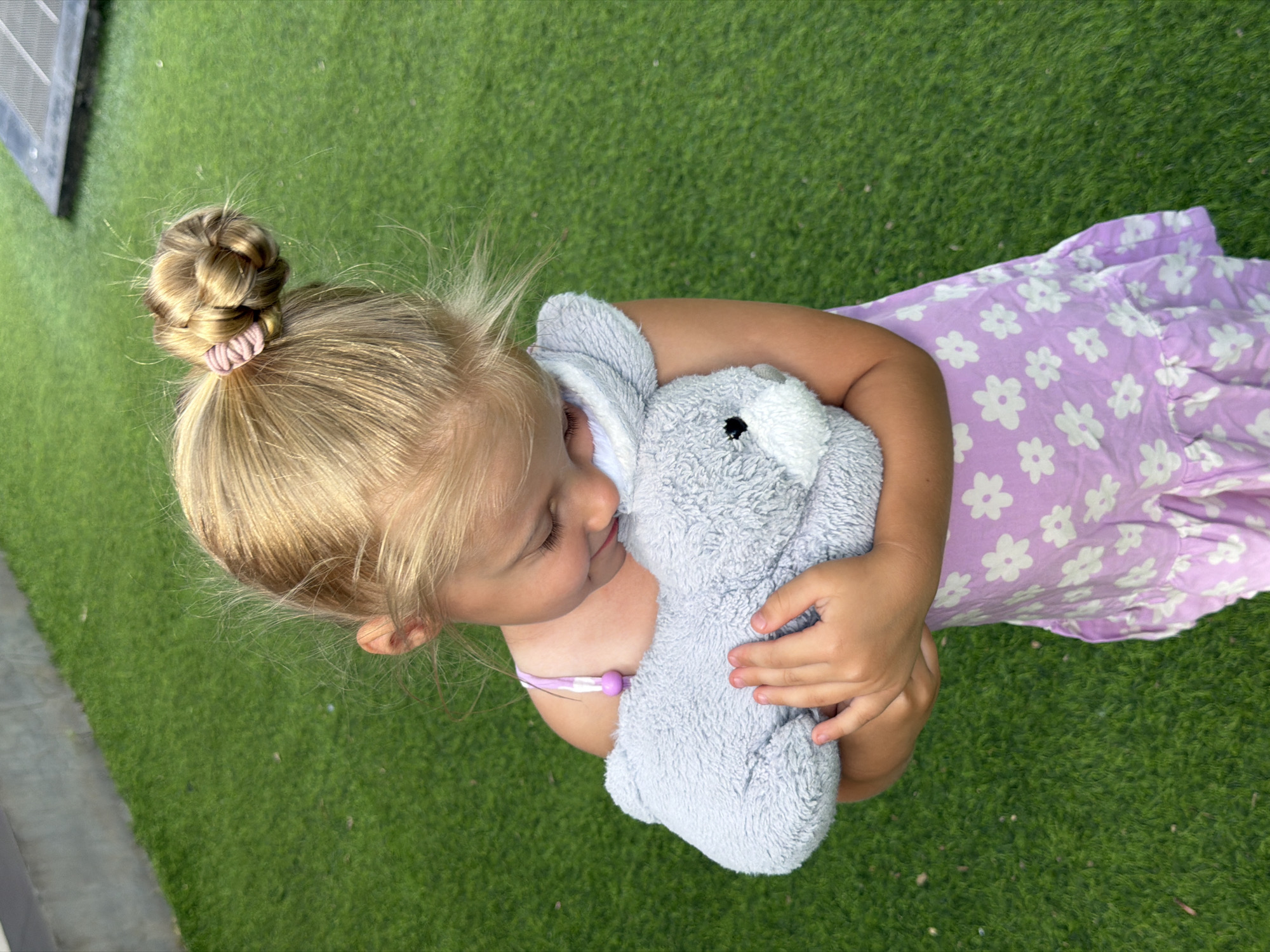 A blonde girl with her hair in a bun holds a gray stuffed bunny. She is wearing a light purple dress with white flowers and a pink and white strap. The girl is looking down at the bunny and smiling. She is standing on green artificial turf. The background is out of focus and appears to be a concrete wall and a dark rectangular object.