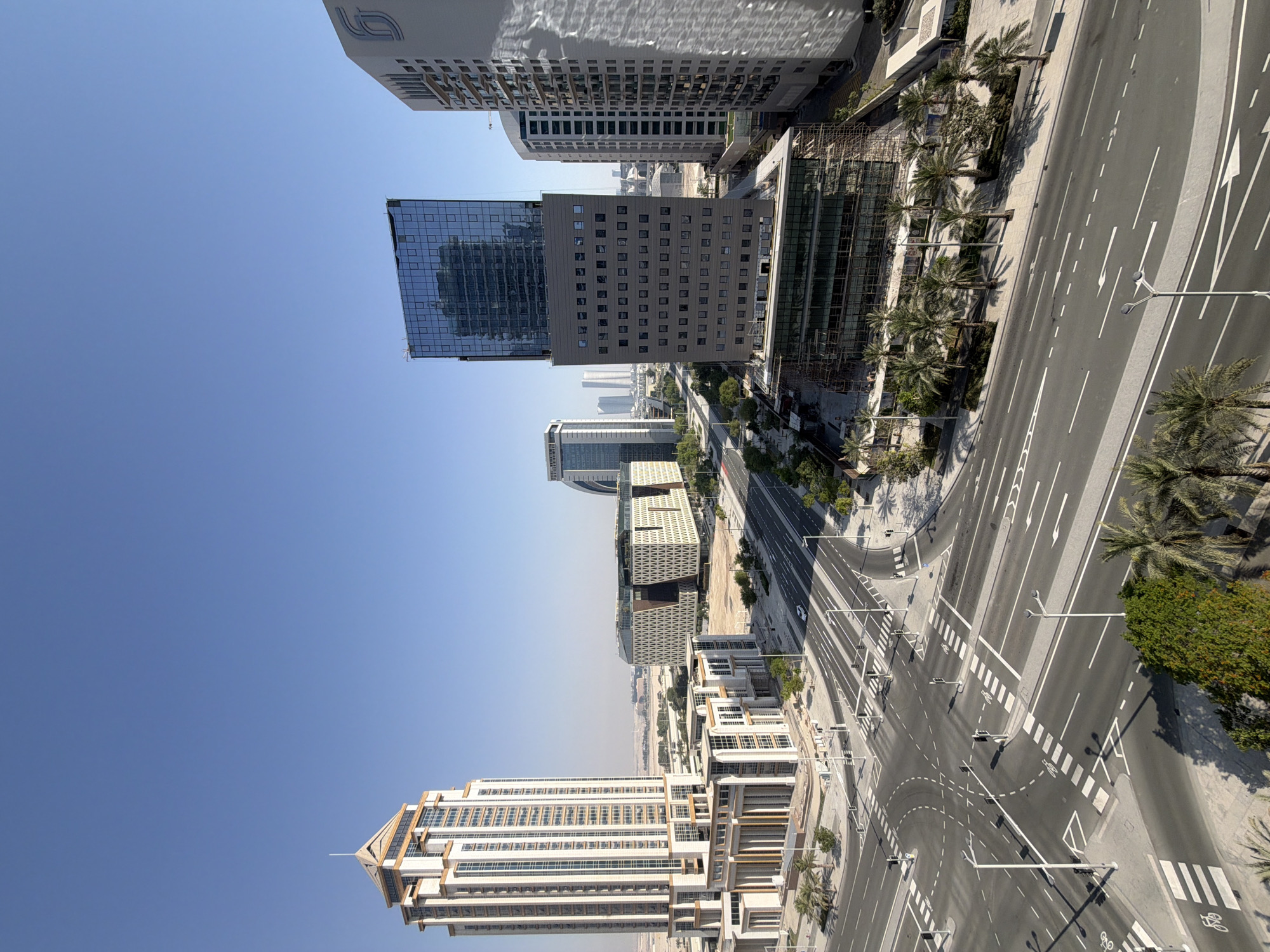A high-angle, wide shot captures a modern cityscape under a clear blue sky. In the foreground, an intersection of multi-lane roads with white dashed lines and pedestrian crossings is visible. Traffic lights and street lamps dot the paved surface. The surrounding area is characterized by low-rise buildings with white facades, palm trees, and sparse vegetation.
Dominating the middle ground are several contemporary high-rise buildings. To the left, a tall, cream-colored building with a pointed roof and numerous windows stands prominently. To its right, a building with a facade of geometric patterns in white and brown is situated. Further to the right, a sleek, dark glass skyscraper with a blue-tinted facade rises, supported by a gray lower section featuring rows of windows. Next to it, another tall, light-colored building with a prominent logo on its facade is visible. In the background, more skyscrapers create a distant urban skyline, and the arid landscape extends to the horizon. One of the buildings in the middle ground appears to be under construction, with scaffolding surrounding its lower floors. A single white car is observable on one of the roads.