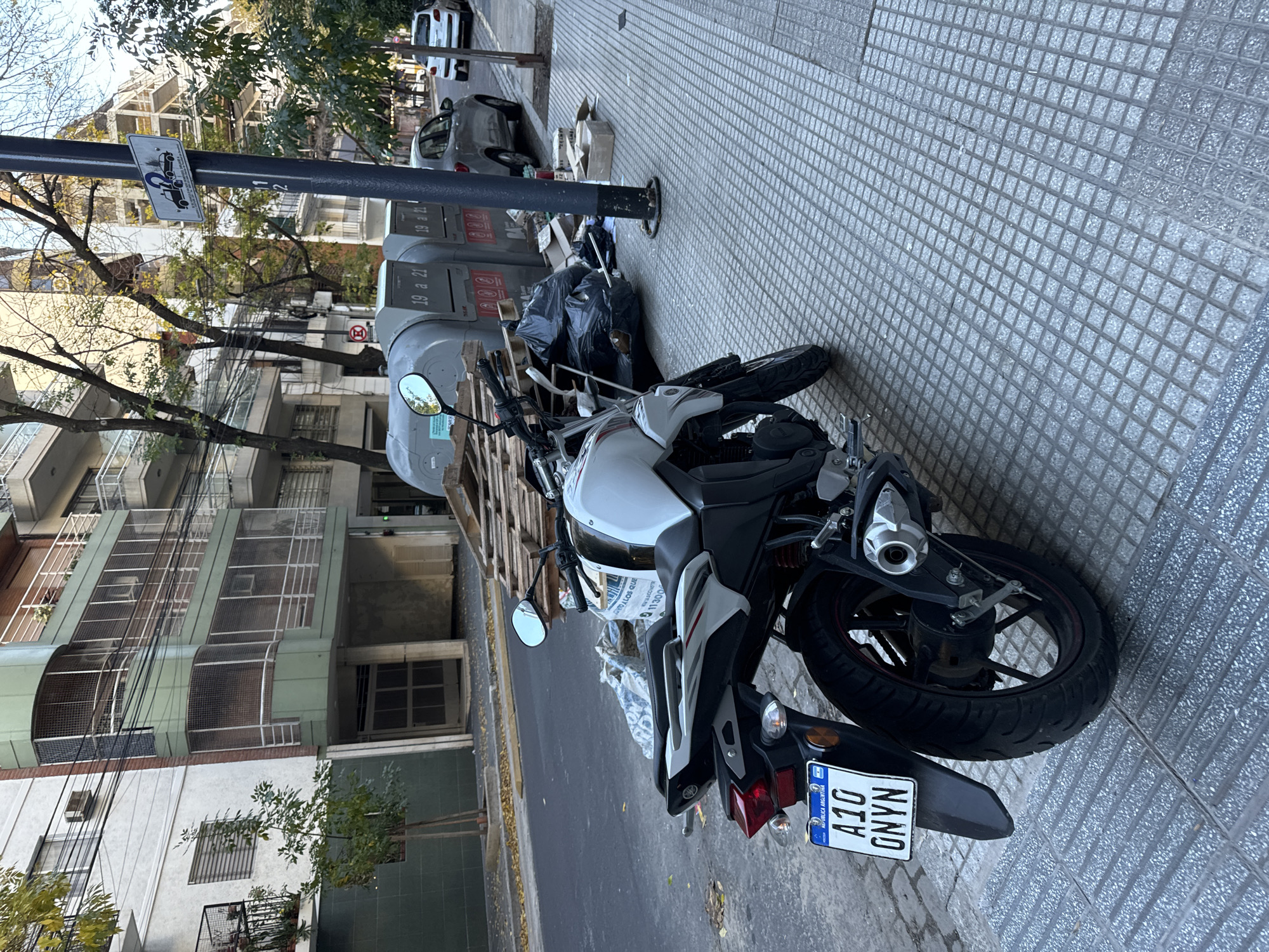 The image presents a motorcycle parked on the sidewalk in a street in Buenos Aires, Argentina. The motorcycle is white and black, with a license plate that reads "A10 ONYN" and includes the text "Republica Argentina". The sidewalk is tiled with a geometric pattern. The motorcycle is positioned near a utility pole. Next to the pole are several large, grey trash containers labeled "19 a 21". Pallets are stacked in front of the trash containers, as well as several black garbage bags. In the background, buildings can be seen on the other side of the street, along with cars parked on the road. Trees are also visible. A street sign is attached to the utility pole. The overall lighting suggests it is daytime.