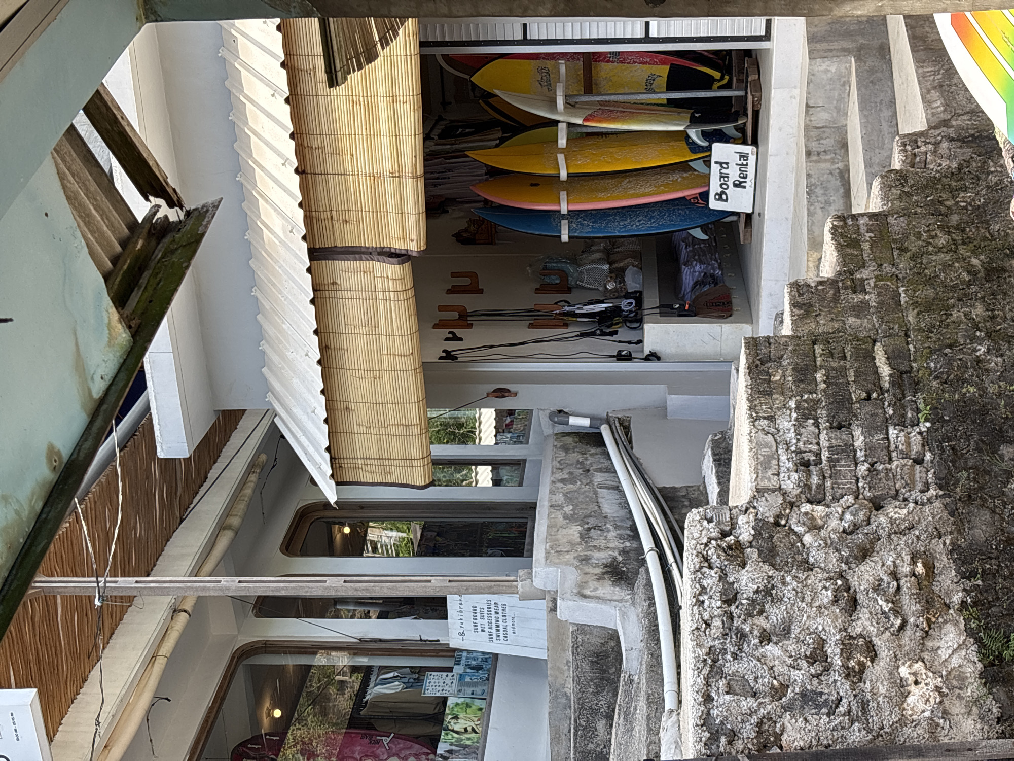 Here is a detailed description of the image:
The image depicts a surf shop in Kuta Selatan, Indonesia, specializing in board rentals and other surf-related merchandise. A prominent sign indicates "Board Rental," and a smaller sign advertises various items, including surfboards, wetsuits, surf accessories, swimming wear, and casual clothing.
Several surfboards of different colors and sizes are displayed on racks inside the shop. Electrical wiring and PVC pipes are visible along the building's exterior near a set of stone steps leading up alongside the building. The building's architecture incorporates a mix of traditional and modern elements, including bamboo blinds, corrugated metal roofing, and weathered stone.