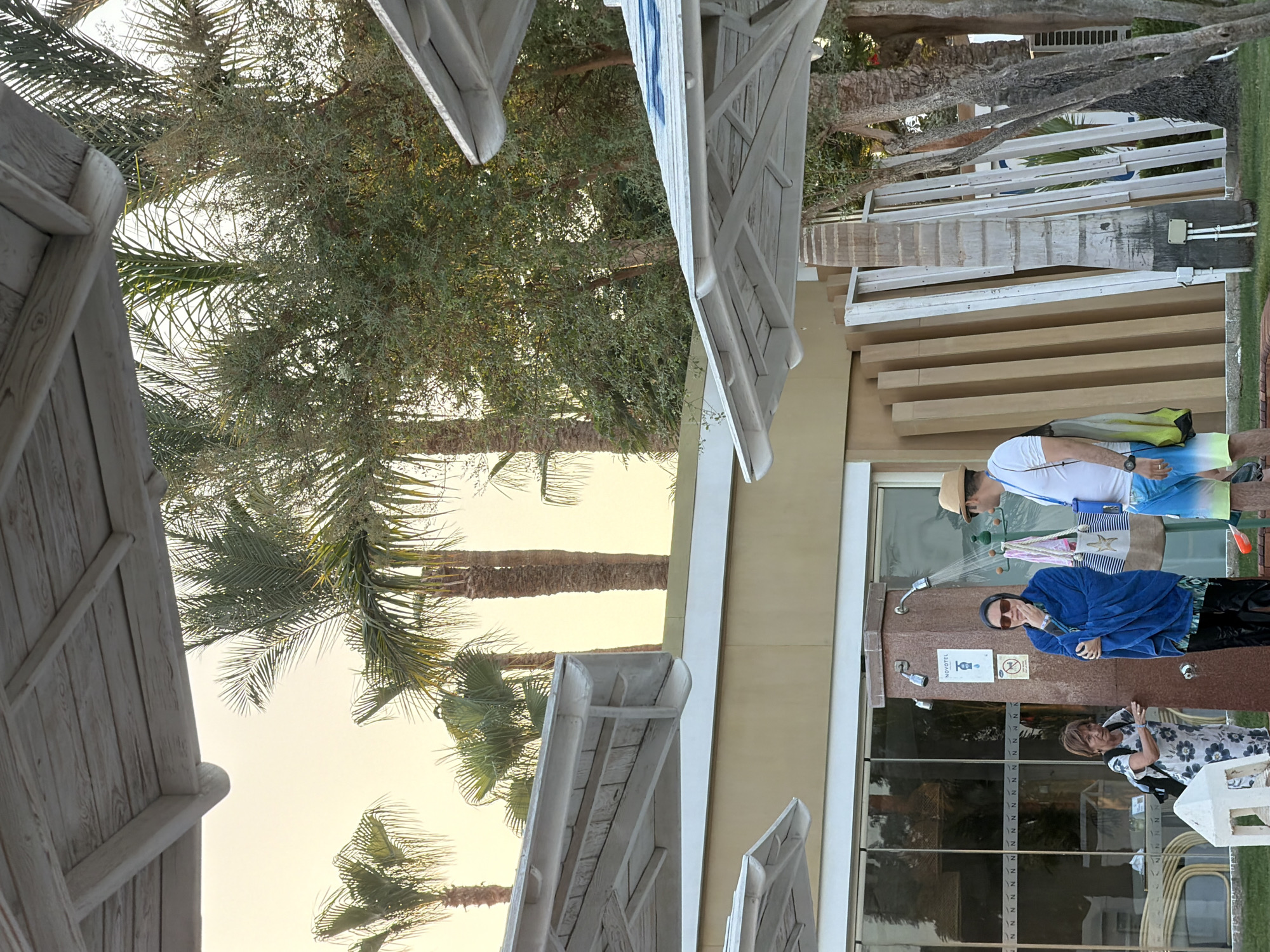 The image depicts an outdoor scene, likely at a resort in Sharm El Sheikh, Egypt. In the foreground, several white wooden structures resembling pergolas or sunshades are visible, with their angled beams and planked roofs casting shadows. Beyond these, a light-colored building with large glass windows forms the backdrop. Palm trees, some with textured trunks, are interspersed throughout the scene, with their fronds reaching towards the sky, which is a soft yellow-orange, suggesting either sunrise or sunset.
In the middle ground, two women and a man are present. One woman, wearing a floral dress and carrying a backpack, gestures towards a shower fixture. Adjacent to her, a woman in a dark hijab and sunglasses is wrapped in a bright blue towel and stands under an active showerhead. A man wearing a hat, white t-shirt, and colorful swim shorts stands to the right of the woman in the blue towel, facing away from the camera. He holds a green bag with a yellow strap. A pink object, possibly a pool float, is on the ground near his feet. Hanging on the shower structure are several bags, including a large tan tote bag with a starfish design.
In the background, leaning against the building are several tall, light-colored wooden planks or frames, suggesting ongoing construction or renovation. The ground appears to be covered in green grass. The overall impression is one of a leisure or resort environment, possibly near a beach or pool area.