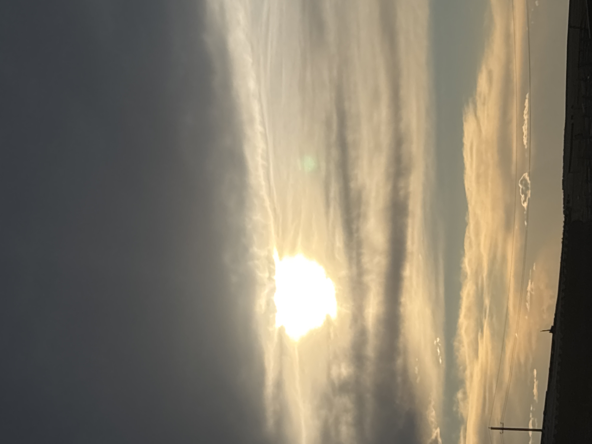 The image shows a view of the sky with the sun shining brightly through the clouds. The sky is a mix of gray and white, with the sun appearing as a bright, undefined shape. In the lower right corner, there are indistinct outlines of what appears to be buildings or structures. There are utility poles and wires visible against the skyline. There is no information to determine the location in the city of Zagatuy, Russia. There are no people or interactions visible in the frame.