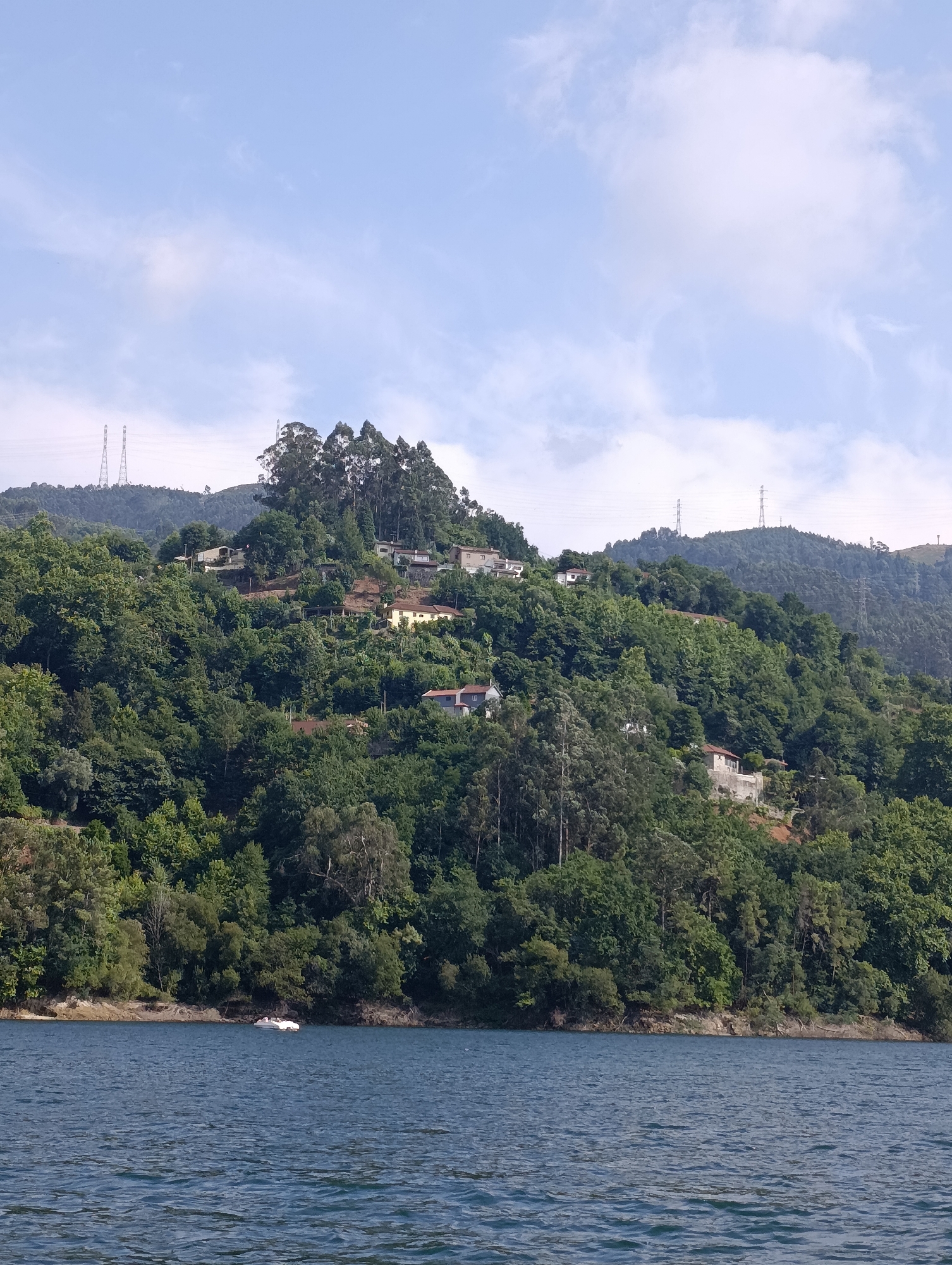 The image shows a view of a body of water with a lush, green hillside in the background. The hillside is covered in dense trees and dotted with houses and structures. Several tall power transmission towers are visible on the distant peaks. The water in the foreground is a deep blue with gentle ripples. A small white boat with people onboard is visible on the water near the shore. The sky is partly cloudy with a light blue hue. This scene is characteristic of the natural beauty found in and around Vieira do Minho, Portugal, a region known for its reservoirs and mountainous landscapes.
