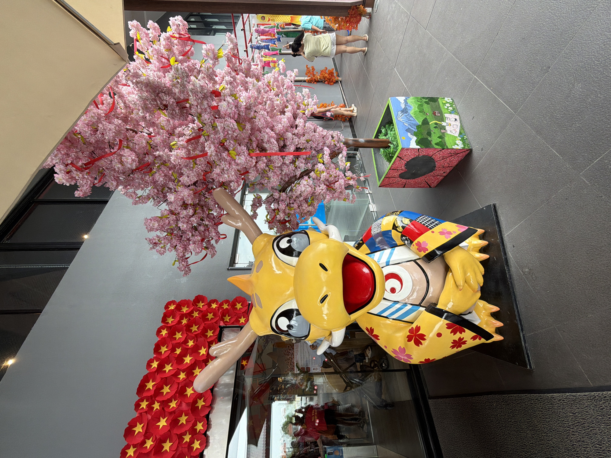 This image depicts an outdoor scene, likely in the city of Đà Nẵng, Vietnam, with decorative elements that suggest a festive or cultural event. In the foreground, a large, brightly colored statue of a yellow dragon stands prominently. The dragon has large, expressive eyes, horns, and is adorned with floral patterns and clothing inspired by traditional Asian designs. To the right of the dragon, a potted artificial cherry blossom tree, covered in pink flowers and red ribbons, adds to the festive atmosphere. The planter itself is decorated with a painted landscape featuring mountains and houses.
In the background, a wall is decorated with a vibrant display of red fabric cutouts shaped like stars. Through a glass entrance to a building, several people can be seen, some appearing to be shopping or browsing. Further down the walkway, another person is visible, taking a photograph with a smartphone, facing away from the camera. The ground is paved with grey tiles. The overall setting appears to be a public space, possibly an entrance to a mall or a decorated street area.