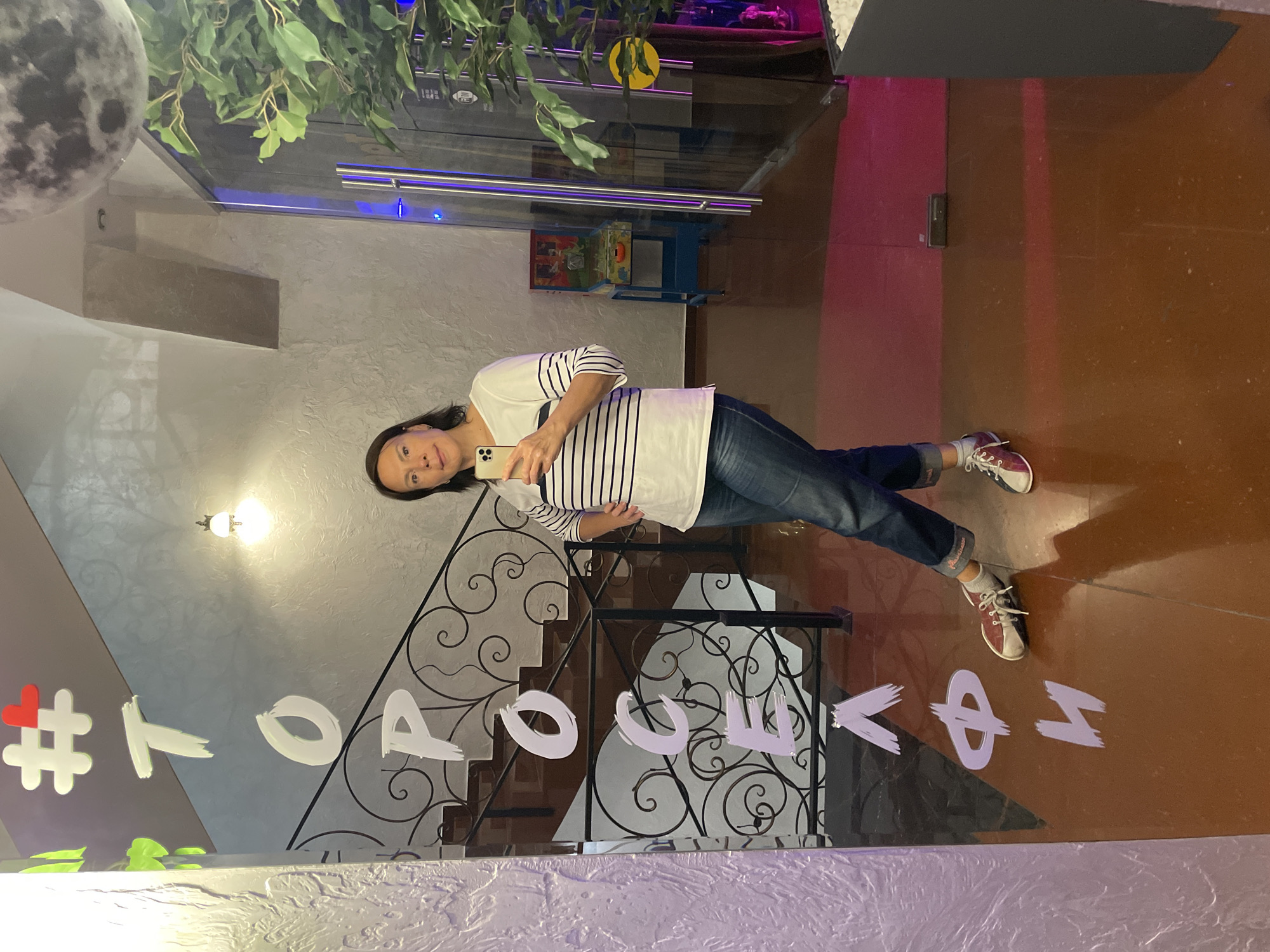A woman in jeans and a striped shirt is standing in a selfie in front of a reflective surface, likely a mirror. The reflection shows a staircase with ornate iron railings and a hashtag message written on the surface. The hashtag "#торосова" is prominently displayed. The background features decorative elements, including a representation of the moon, plants, and unique lighting fixtures. A glimpse of a hallway with a pinball machine suggests the interior of a recreational or entertainment facility. The location is in Sochi, Russia, evidenced by the Cyrillic text in the hashtag.