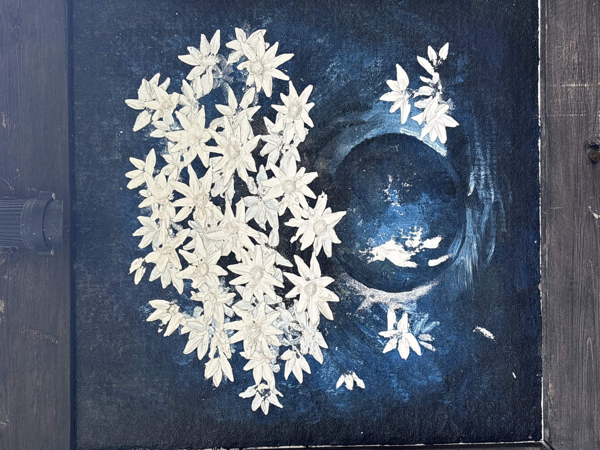 A painting depicts a bouquet of white, star-shaped flowers in a dark, round vase against a deep blue, textured background. The flowers are rendered with thick impasto, giving them a raised, sculptural quality. Some loose flowers and petals are scattered around the vase and in the foreground. The background has subtle variations in blue tones, with some lighter, brushstroke-like textures that suggest light or movement. A dark wooden frame surrounds the canvas. The overall impression is one of artistic expression, with no discernible elements that place it in a specific location in Jiyang District, China, or any other city.