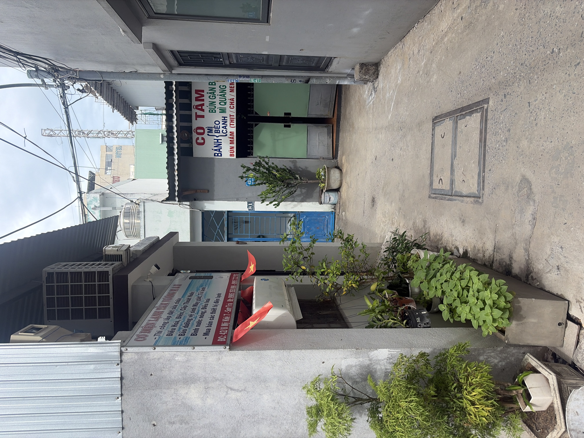 This is a medium-shot, eye-level, outdoor photograph taken on a bright, overcast day in a narrow alleyway in the Sơn Trà district of Đà Nẵng, Vietnam. The alleyway appears to be a commercial area, with buildings on either side.
On the left, a building with a corrugated metal roof and grey concrete walls is visible. A sign with Vietnamese text, listing services related to air conditioning repair and sales, is mounted on the wall. Two red Vietnamese flags with yellow stars are attached to the wall above a white air conditioning unit. A potted green plant with small, serrated leaves stands in the foreground, to the left of the building.
In the center of the alleyway, a blue door is slightly ajar. Beyond the door, a glimpse of a staircase can be seen. Further down the alley, a building with a sign that reads "CÔ TẨM" (which translates to "Mrs. Tam's") is visible. This sign advertises food items such as "CƠM TẤM" (broken rice), "BÁNH BÈO" (steamed rice cakes), "BÚN GÀN BÁNH CANH" (noodle soup with chicken and thick noodles), and "MÌ QUẢNG" (Quang-style noodles). Two large, pale green double doors lead into this establishment. A small, spiky green plant is potted in front of this building.
The ground of the alleyway is paved with concrete, with some patches of darker, exposed aggregate concrete. In the center of the alley, a rectangular metal grate is set into the pavement. The right side of the alley is lined with a grey wall, with a dark-colored window and a vertical metal pole with numerous electrical wires running overhead. The overall scene depicts a typical urban alleyway in a Vietnamese city, suggesting a mix of residential and small business activity.
