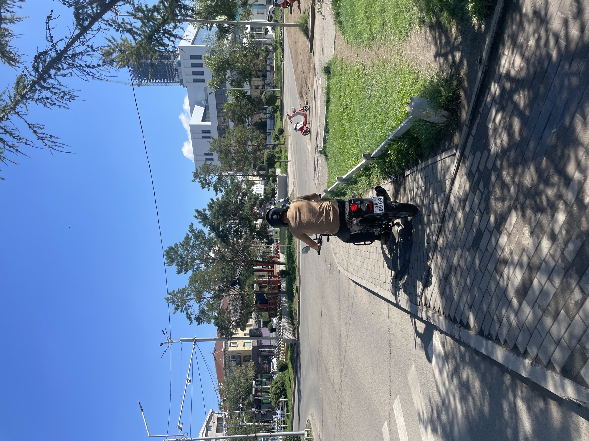 The image captures a sunny day scene in Ulaanbaatar, Mongolia, specifically near a street intersection. A person is riding a motorcycle on a paved sidewalk, facing away from the camera. The rider is wearing a helmet and a light brown shirt. The motorcycle has a license plate visible, reading "YK 09 69". In the background, there's a paved road with a pedestrian crossing. Beyond the road, there are buildings, including a modern white building with multiple windows and a construction site with a crane. Traditional-style structures with red roofs and pavilions are also visible, suggesting a mix of architectural styles. Green trees and grassy areas are interspersed throughout the urban landscape, casting shadows on the ground. A red scooter is parked on the side of the road in the mid-ground. The sky is clear blue.