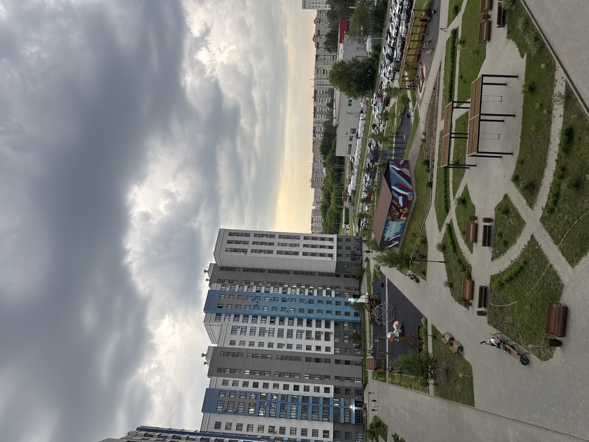 An aerial, oblique view captures a modern residential complex in Barnaul, Russia, under a cloudy sky. The foreground features a children's playground with swings, a climbing structure, and a sandpit. Paved pathways wind through manicured green spaces dotted with wooden benches and trash receptacles. To the left, a multi-story apartment building with a facade of grey, white, and blue panels dominates the scene. In the midground, a parking lot is filled with numerous cars, and a long, low-rise building with a white facade is visible. Beyond this, a dense array of similar apartment buildings stretches across the horizon, interspersed with trees. The sky is overcast with dramatic grey clouds, with a sliver of warmer light breaking through near the horizon. Several people are visible in the image, including a few walking in the distance, a couple sitting on a bench, and a child playing near the sandpit.