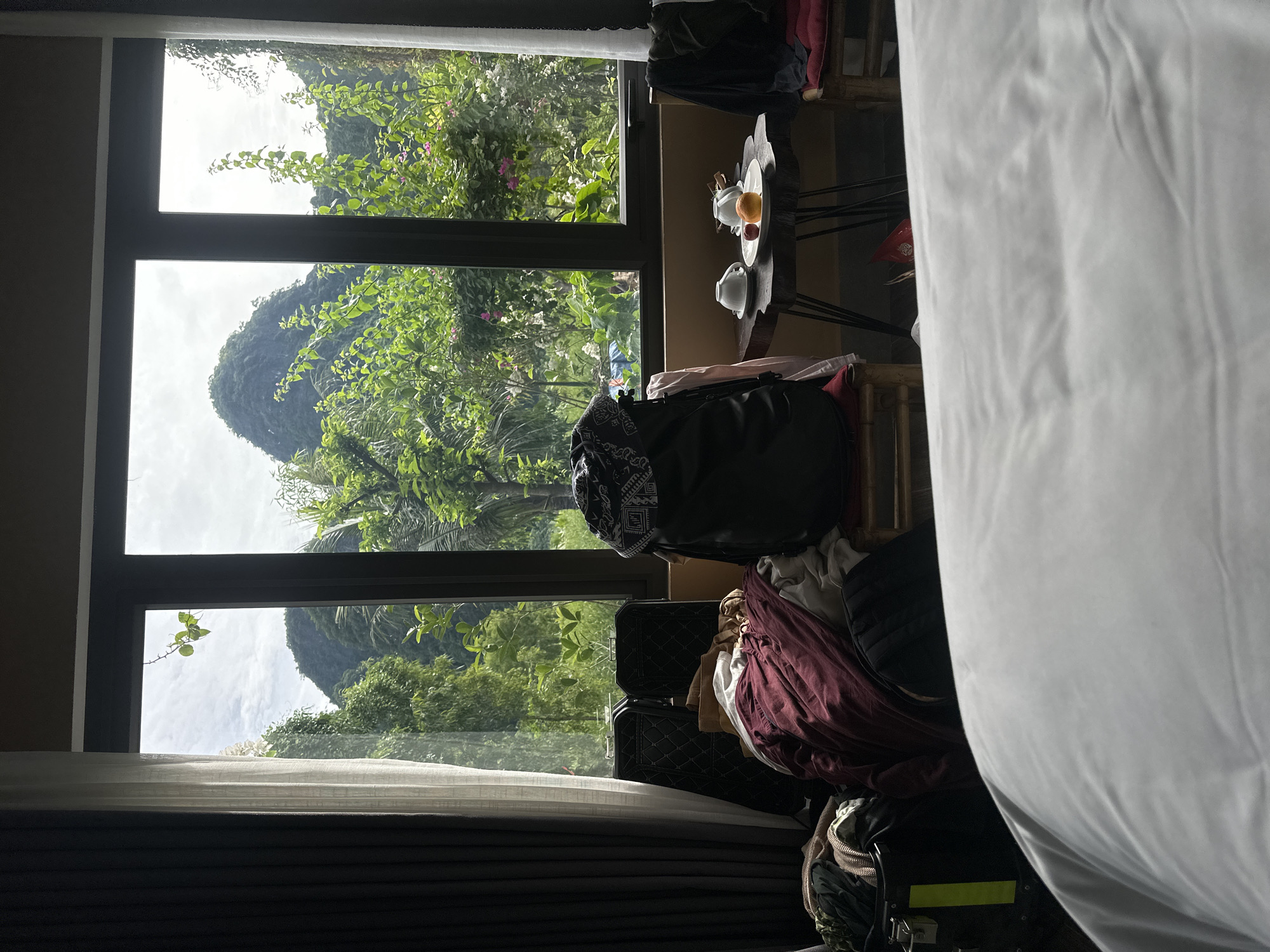 The image depicts a cluttered hotel room with a large window offering a view of lush green mountains and vegetation. In the foreground, a white bedsheet fills the bottom portion of the frame. To the left, dark curtains hang beside the window. Near the window, several pieces of luggage and piles of clothing are visible, including a black backpack and a burgundy garment. A small wooden table stands in front of the window, holding a white teapot, a cup and saucer, and fruit, including an orange and a red apple. Two chairs, one partially visible with a red cushion, are positioned near the table. The background reveals a dramatic mountainous landscape with steep, tree-covered slopes under a cloudy sky. The scene suggests a travel lodging setting in a natural environment, likely in the vicinity of Nam Hoa Lư Ward, Vietnam, known for its karst topography.