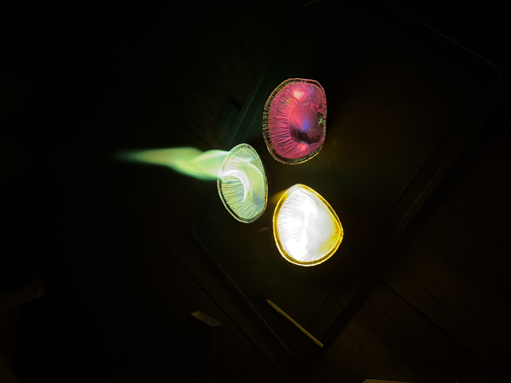 The image shows three colorful light fixtures against a dark background, possibly a ceiling. The light fixtures are arranged in a triangular pattern. The top fixture is pink/red, the fixture on the left has a green streak originating from it, and the bottom fixture is white. The background appears to be a dark wood surface with some linear features suggesting paneling or planks. The image was taken in a dimly lit environment. There is no indication of the location being in Chaikovsky, Russia.