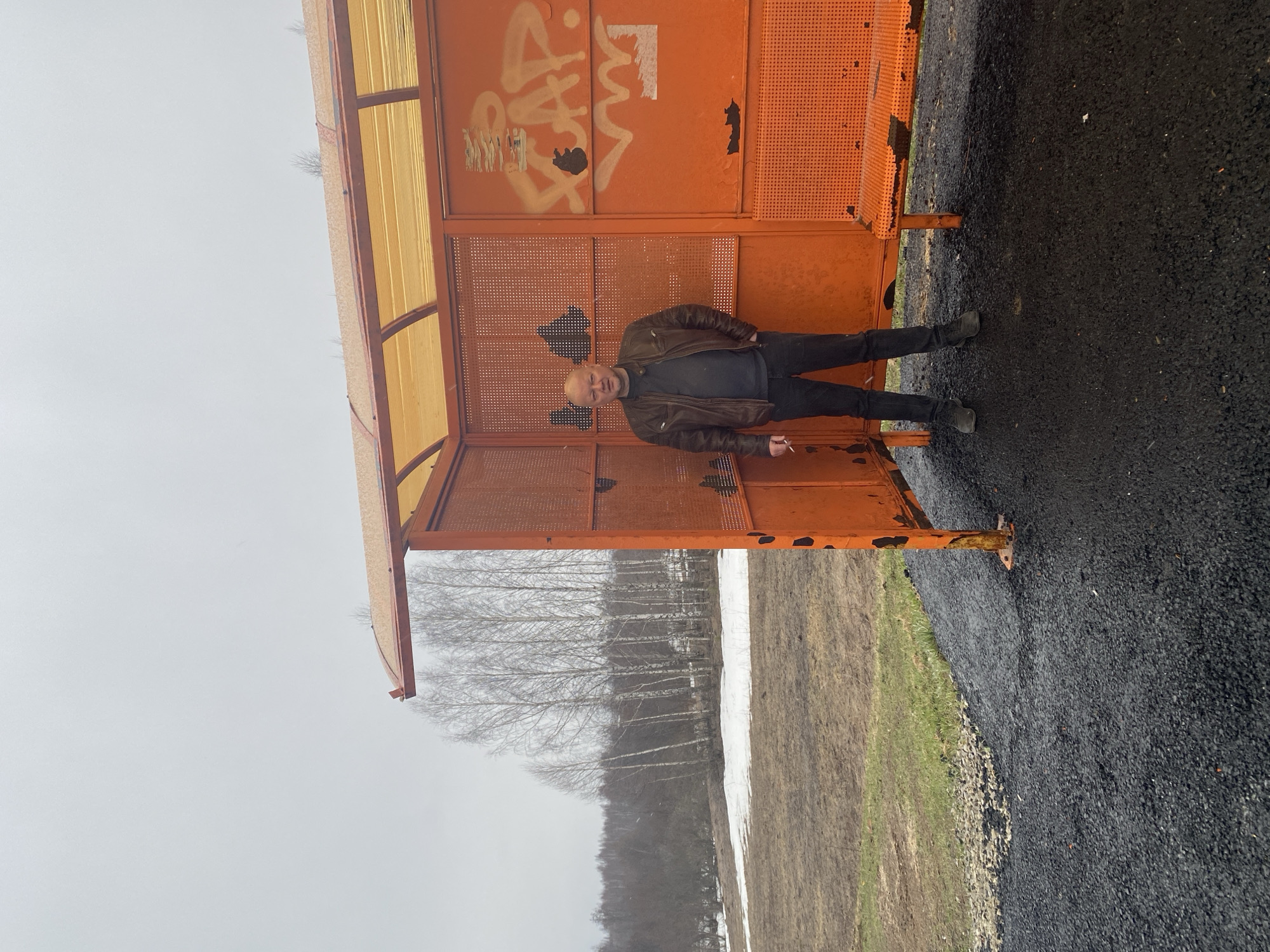 A middle-aged man is standing inside a rusted orange structure with a translucent yellow roof. He is wearing a brown leather jacket, a dark shirt, and dark pants. The structure is located in a landscape with patches of snow and bare trees in the background. The ground is a dark, paved surface. The city is Буль-Кайпаново, Russia. There is graffiti on the orange structure behind the man.