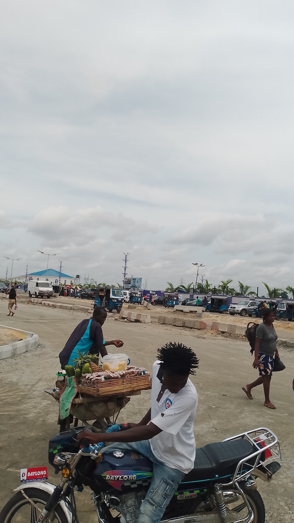 The image shows a scene in what appears to be an urban setting with an overcast sky. In the foreground, a man sits on a blue Daylong motorcycle. Another man stands behind him, tending to a wooden cart loaded with fruits, including pineapple and what appears to be meat skewers. A woman with a backpack walks by in the background to the right, and other people can be seen further in the background. A variety of vehicles, including vans and auto-rickshaws, are parked along the street, with lampposts and trees visible in the distance. Concrete barriers are in the middle of the street, indicating construction or road management.