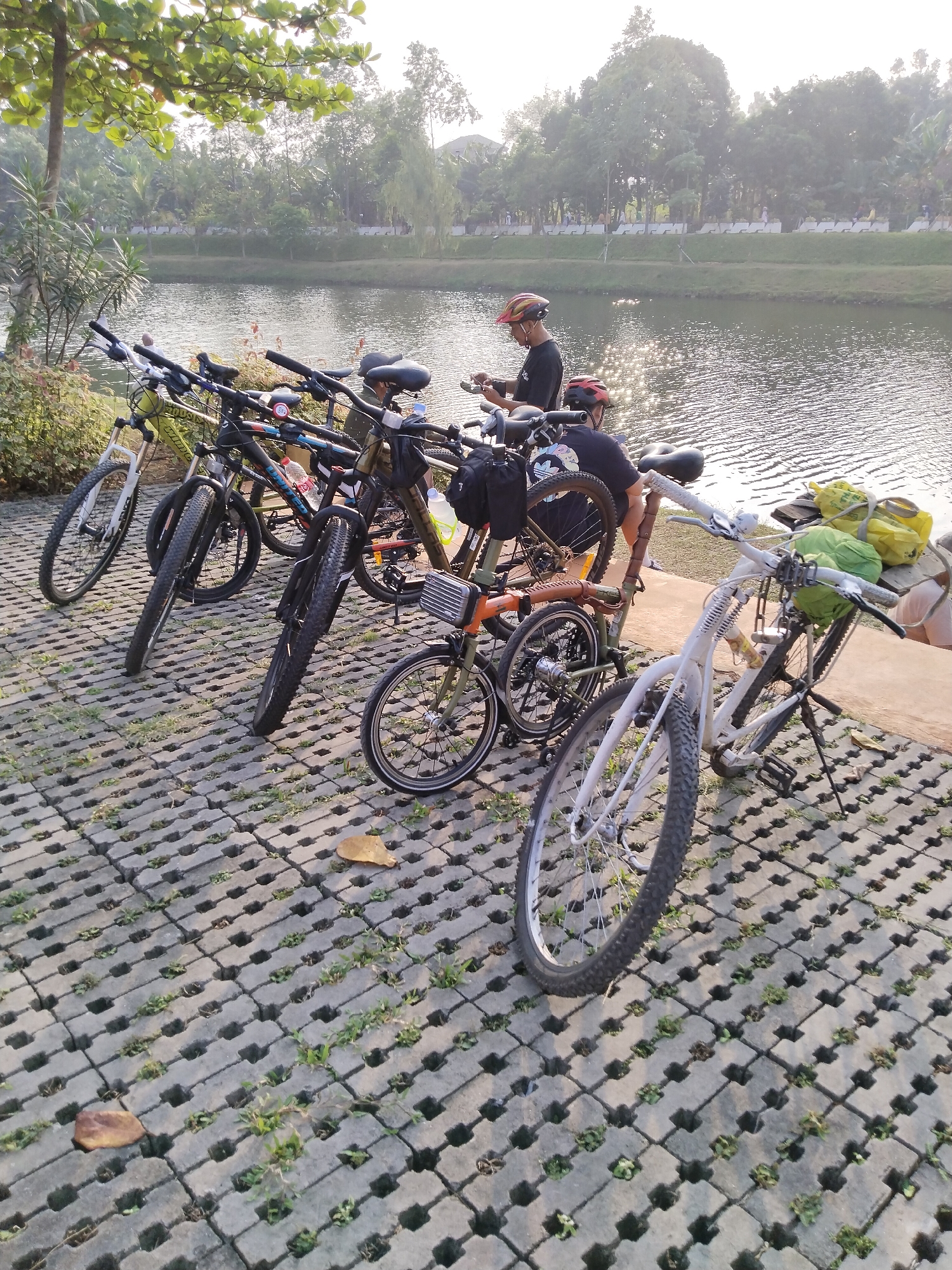 In South Jakarta, Indonesia, a group of bicycles is parked on a brick-paved area next to a body of water, likely a lake or river. The scene appears to be outdoors in a park or recreational area, with trees lining the opposite bank of the water. The bicycles are of various types, including what appear to be mountain bikes and folding bikes, some with bags or backpacks attached. Two individuals, wearing cycling helmets, are standing near some of the bicycles, possibly preparing for or finishing a ride. One person is looking down at something in their hands, while the other is partially obscured. The lighting suggests it is either early morning or late afternoon, with soft sunlight filtering through the trees. The overall atmosphere is one of casual outdoor activity and recreation in a semi-urban natural setting.