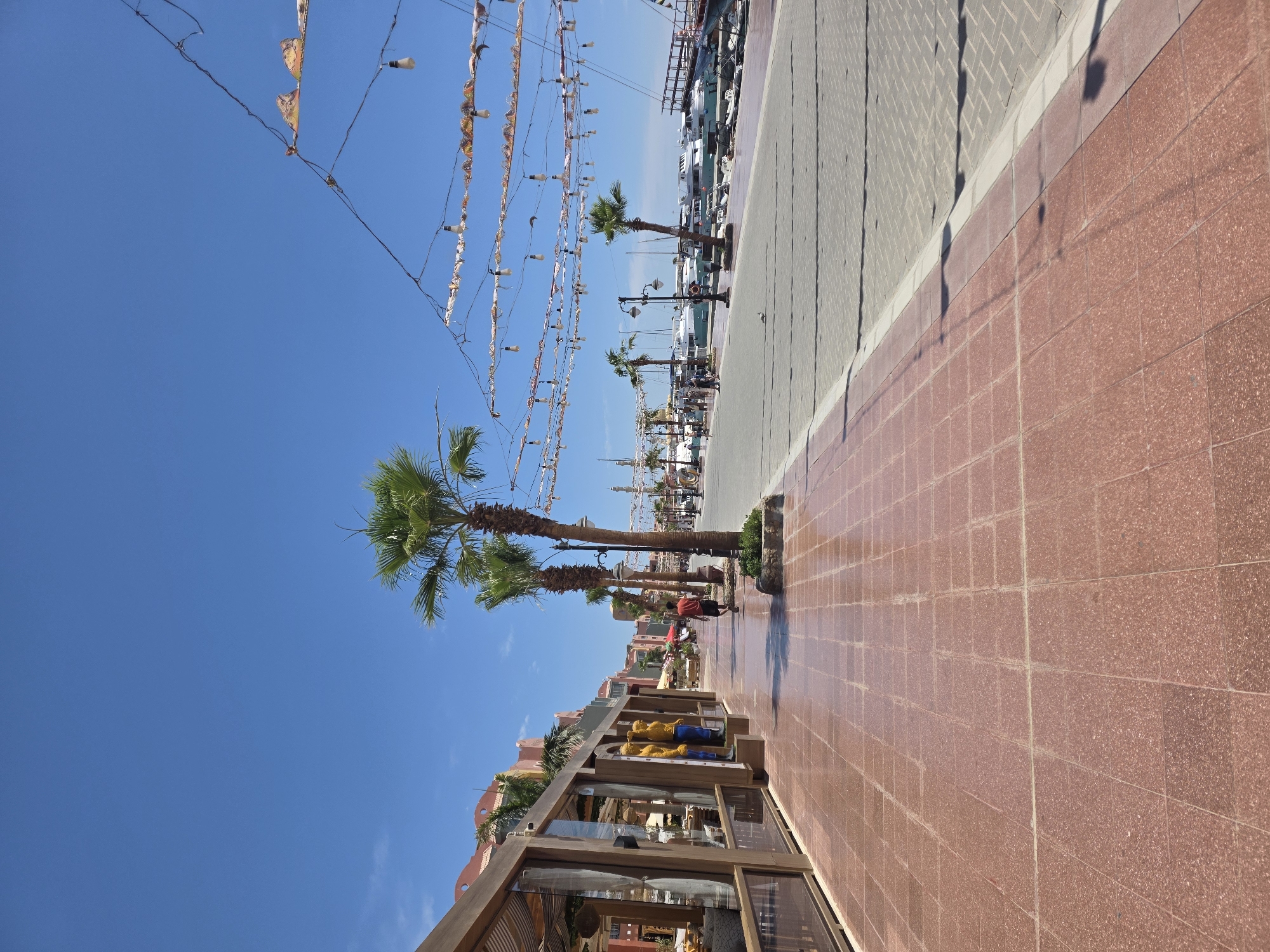 The scene is a promenade in Hurghada, Egypt, viewed from a slightly elevated perspective, looking lengthwise along a wide pedestrian walkway paved with red tiles. A parallel walkway on the right is paved with grey bricks. Palm trees line the walkway. Decorative flags and lights are suspended above. A building with glass-fronted shops is on the left. Several boats are visible in the background beyond the palm trees. A few people walk along the walkway. The sky is clear and blue.