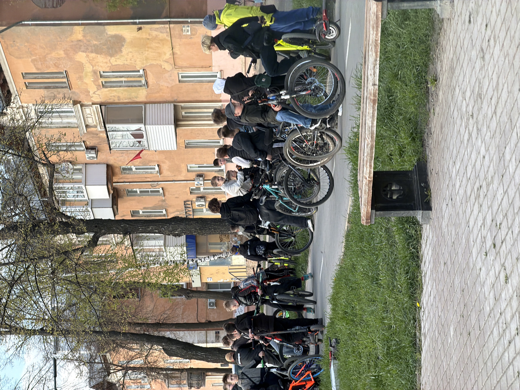 A group of people riding bicycles, possibly a class of students, is positioned in a line. The scene appears to be in Votkinsk, Russia. The buildings in the background are multi-story apartment buildings. The trees are just starting to grow leaves. A paved sidewalk is visible, as is a narrow strip of grass with a park bench. The group of people is riding on the street beside the sidewalk. The people are wearing jackets.