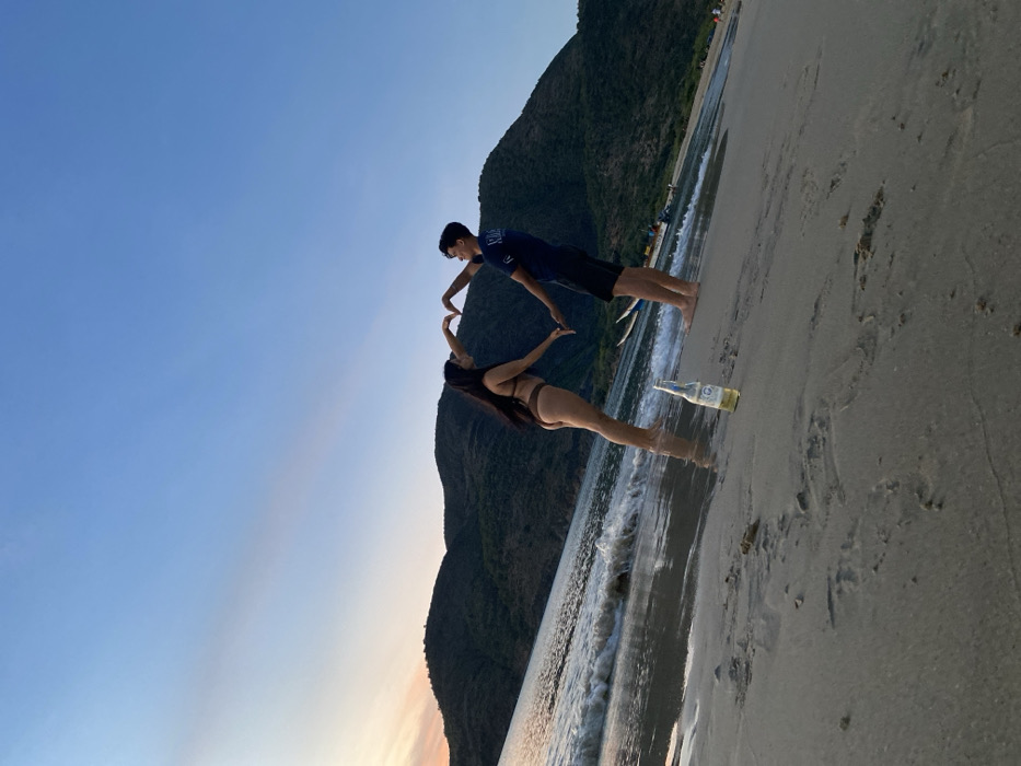 A man and woman stand on a beach, appearing to touch fingertips to form a heart shape. The woman wears a bikini, while the man is in a t-shirt and shorts. Mountains loom in the background under a blue sky. A bottle sits on the sand near the ocean. The beach is mostly empty, and the image is taken at an angle.
