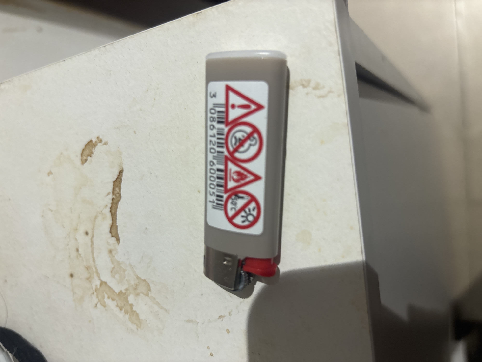 A disposable lighter rests on a stained white surface, possibly a table. The lighter is gray with a red lever and features a sticker with warning symbols and a barcode. The symbols include a circle with a diagonal line through a sun icon, indicating to avoid direct sunlight, and a triangle with an exclamation mark, signifying a general warning. The barcode appears above the symbols, followed by numbers that are partially obscured. The surface beneath the lighter exhibits brown stains, suggestive of spilled liquid or food. The lighting in the scene is dim, casting a shadow to the left of the lighter.