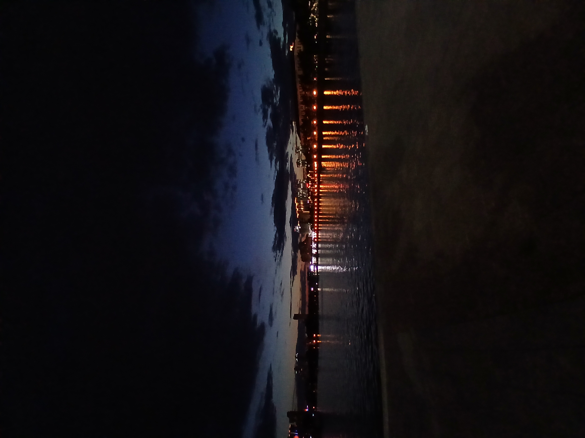 The image captures a waterfront view of Yekaterinburg, Russia, at dusk. A body of water, likely the Iset River, occupies the lower half of the frame, with its surface reflecting the lights of the city skyline. The distant skyline is illuminated by numerous lights, suggesting buildings and possibly structures like bridges or embankments. The sky above is a deep, dark blue, transitioning to a lighter hue near the horizon, indicating the fading daylight. Dark clouds are visible in the upper portion of the sky, adding to the dramatic twilight atmosphere. The foreground is dark and out of focus, suggesting it might be a platform or embankment from which the photograph was taken. There are no people or vehicles visible in the image. The scene is tranquil and conveys the ambiance of a city at nightfall.