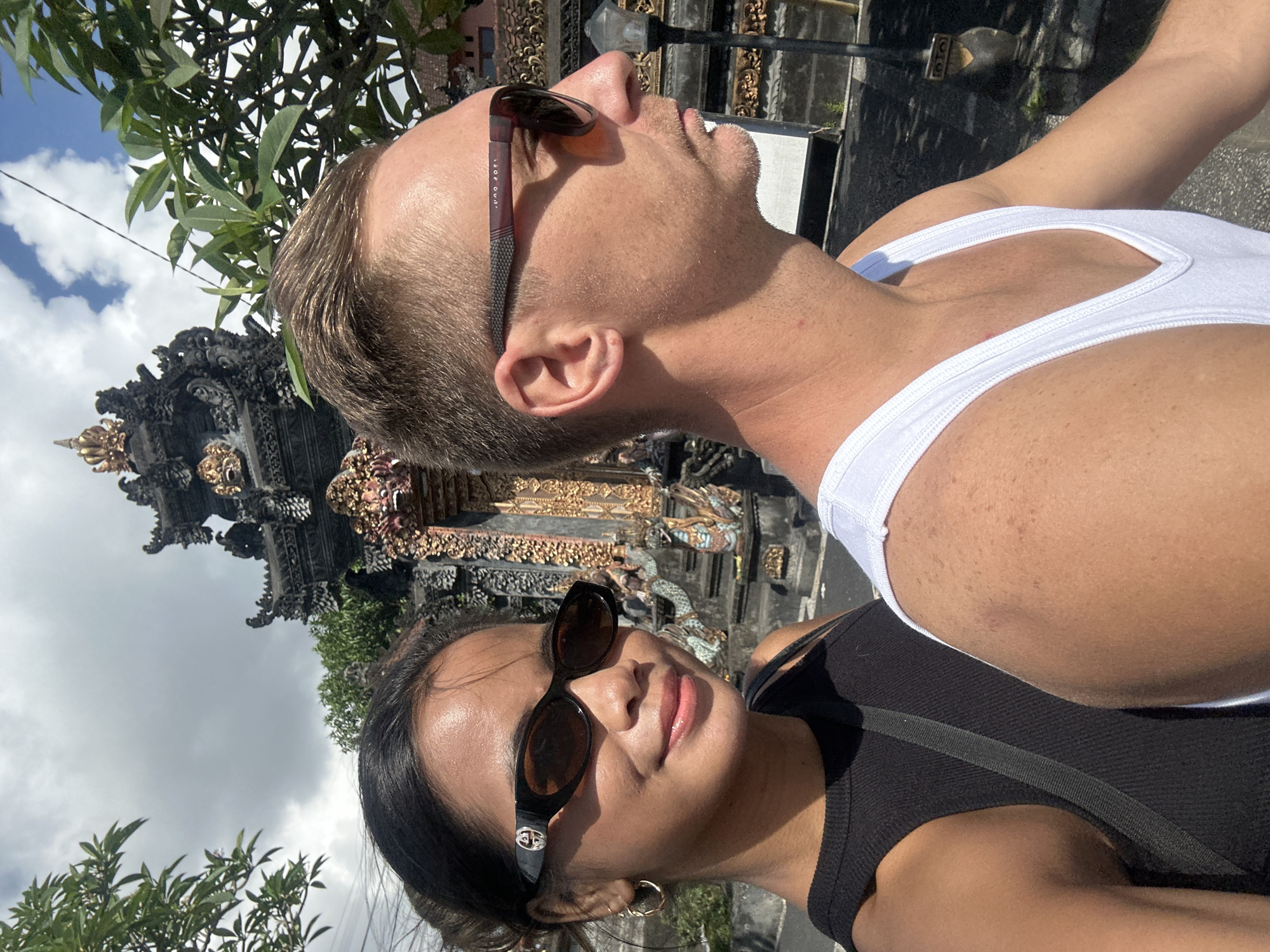 A medium shot captures a man and a woman in the foreground, with a decorative Balinese temple gate in the background, suggesting a location in Sukawati, Indonesia. The woman, on the left, is looking towards the camera with a slight smile. She has dark hair pulled back and is wearing a black tank top and stylish dark sunglasses with a prominent "GG" logo on the side. A gold hoop earring is visible on her left ear. The man, on the right, is in profile, looking to his right. He has short, light brown hair and is wearing a white tank top and dark sunglasses. His skin appears to be sun-kissed, with some freckles visible. The temple gate behind them is intricately carved with dark stone and adorned with gold accents and colorful decorative elements, including what appears to be a statue of a mythical creature. Lush green foliage frames the scene from the top left and right, and the sky above is partly cloudy. The overall lighting suggests it is daytime, likely mid-morning or afternoon.