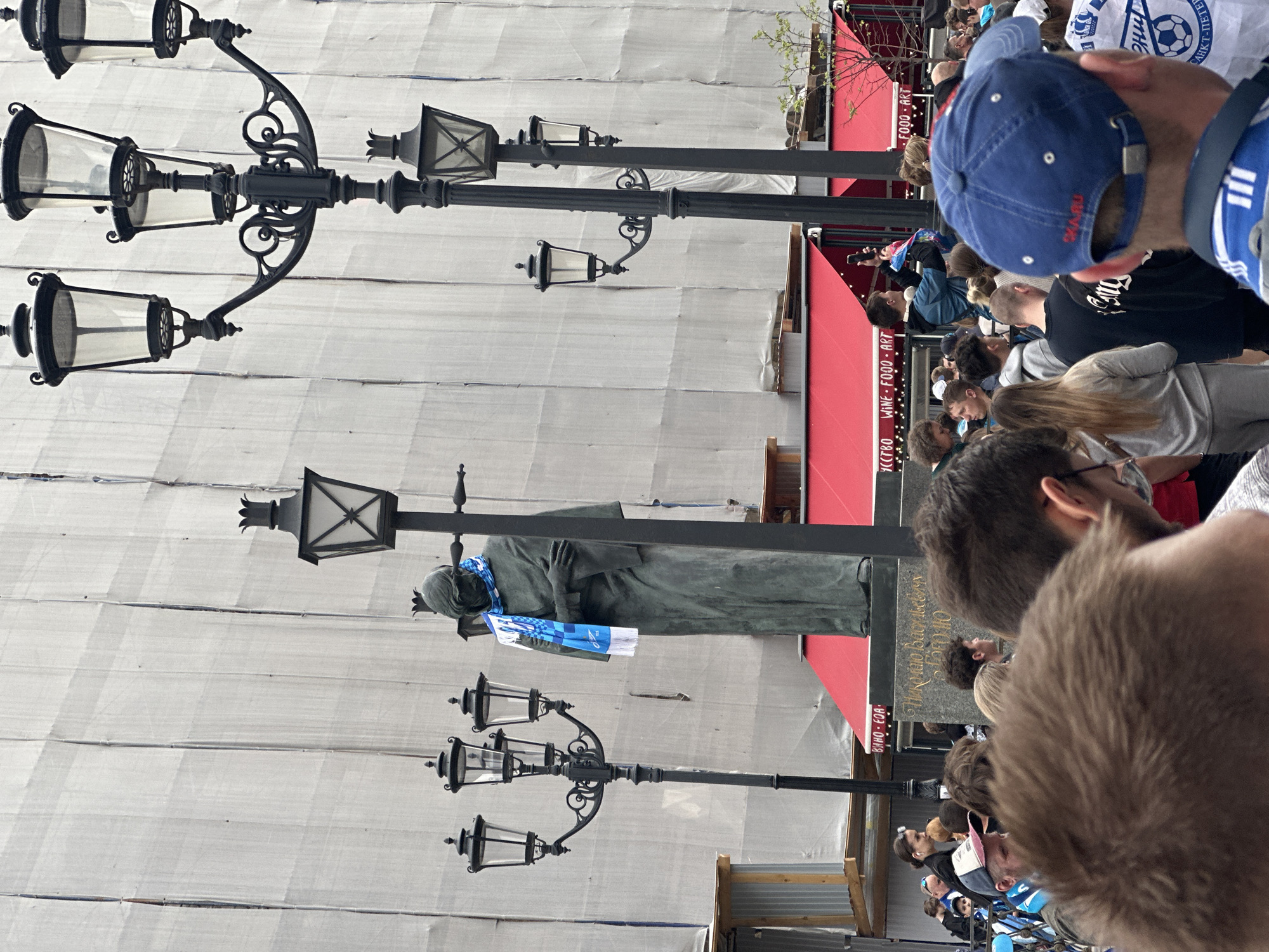 This outdoor shot depicts a crowd of people gathered near a statue in Saint Petersburg, Russia. The statue appears to be of a historical figure, draped with a blue and white scarf. The backdrop includes buildings with red awnings and signs in Russian and English (e.g., "WINE FOOD ART"). Several ornate black lampposts are visible, some with multiple lamps. There are indications of construction or renovation work, with a white tarp or covering present in the background.