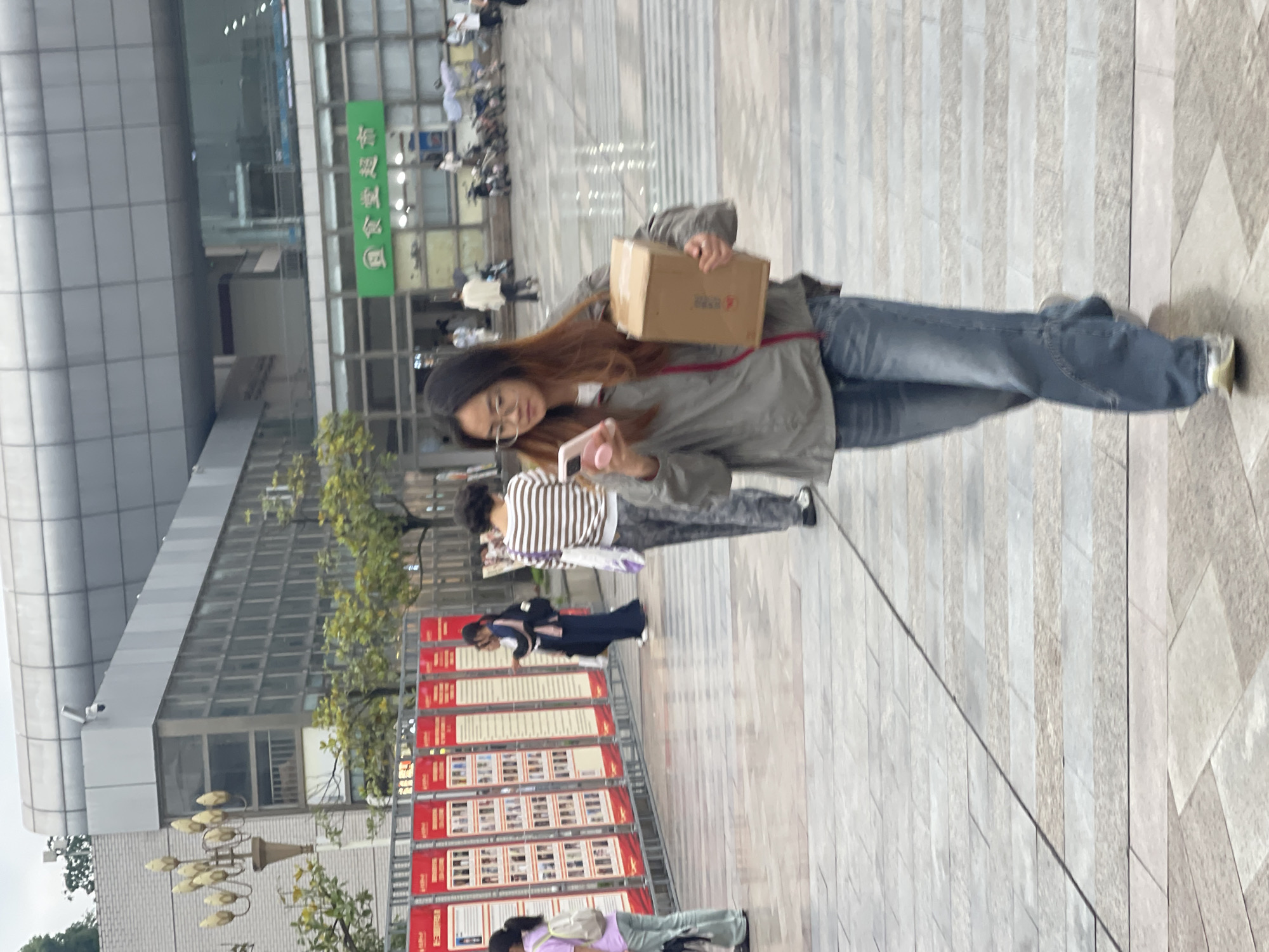 The image shows a woman standing on a paved outdoor area, holding a cardboard box. She wears a gray jacket, blue jeans, and a black beanie, and she appears to be looking at a pink mobile phone. Behind her, other people walk along the pavement. Red poster boards stand to the left, featuring rows of pictures. In the background, a large building with rows of windows is visible. Based on the visible signage, the location is likely in the Chongqing High-tech Zone, China.