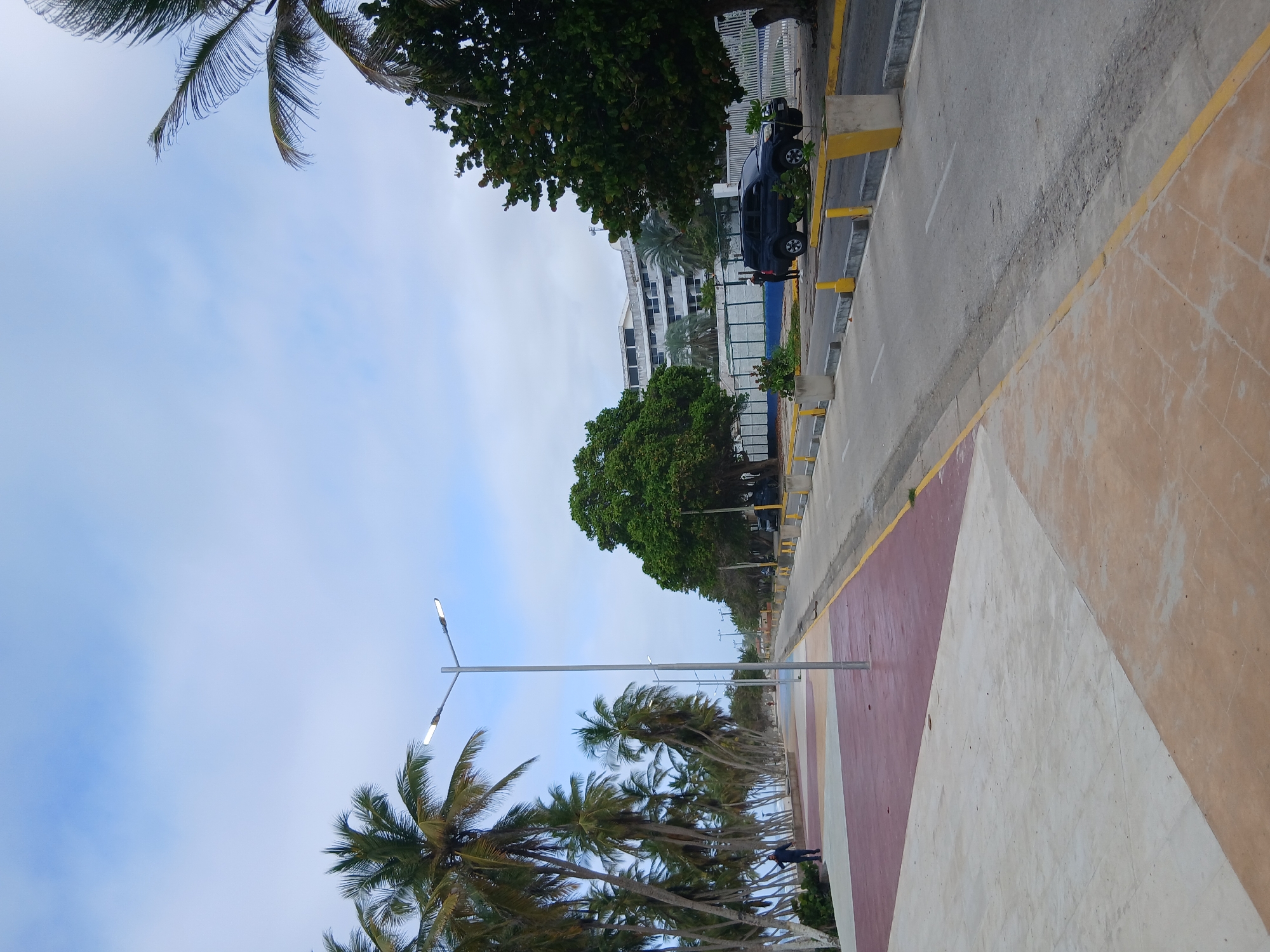The image depicts a scenic view of a coastal walkway and road at Playa El Agua, Venezuela. The walkway features a modern design with geometric sections of light beige, reddish-brown, and a lighter tan color. A tall, silver lamppost with two lights stands prominently on the walkway. To the left, a line of lush green palm trees stretches along the coastline, with a person in a dark jacket walking away from the camera on the walkway.
Adjacent to the walkway is a grey asphalt road, bordered by yellow and grey concrete planters. A dark-colored SUV is parked on the right side of the road. In the background, a large, multi-story white building with a curved facade is visible, flanked by mature green trees. The sky is partly cloudy, with a mix of blue and white clouds. The overall scene suggests a well-maintained public space designed for leisure and transit in a tropical coastal environment.