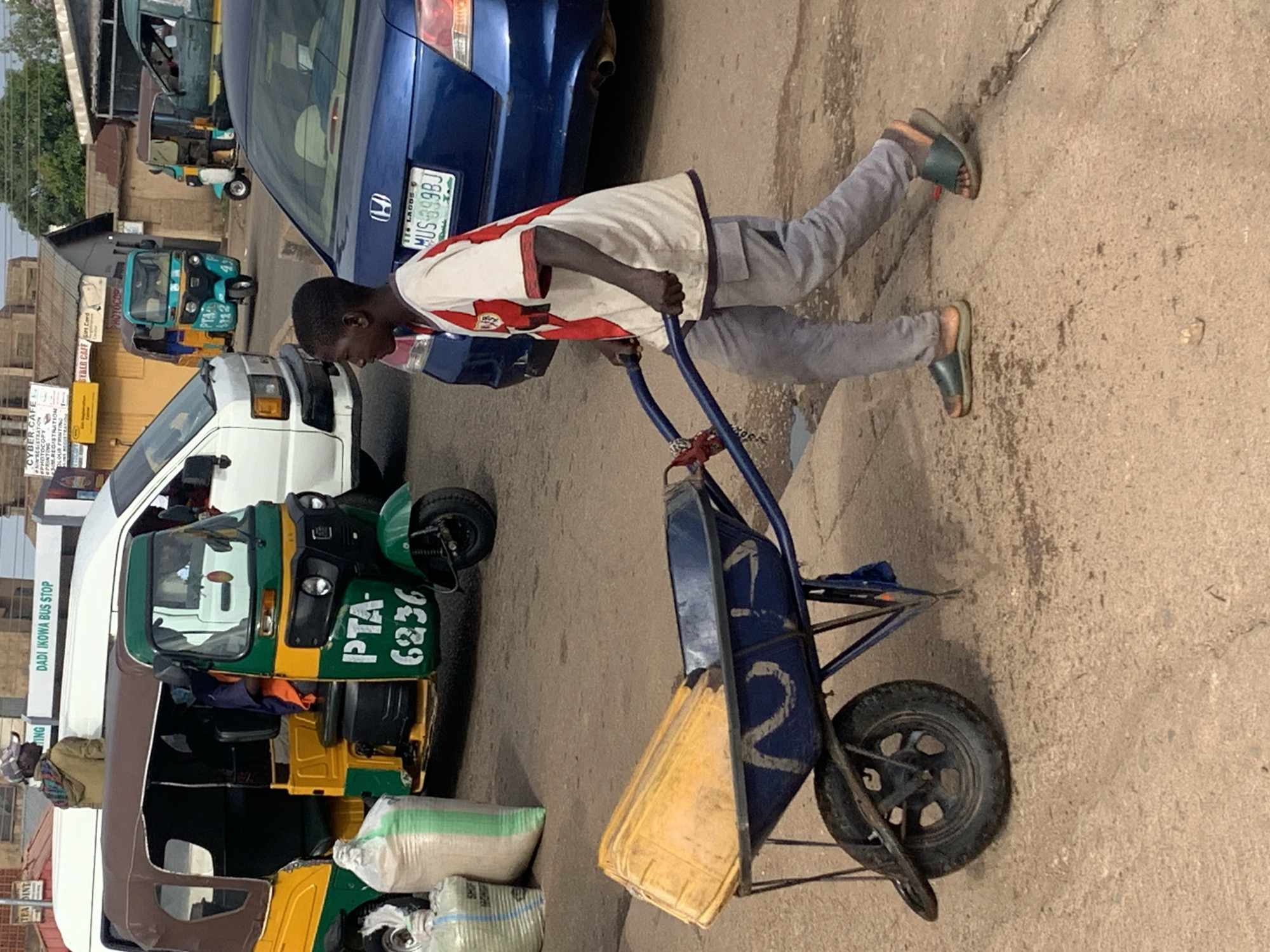 A young boy, wearing a white and red shirt and grey pants, pushes a blue wheelbarrow loaded with yellow containers through a street in Gura Top, Nigeria. The boy is walking to the right of the frame. In the background, a green and yellow auto-rickshaw is parked next to a white van. Several buildings and a blue car are also visible. The ground is paved with dirt and some debris.