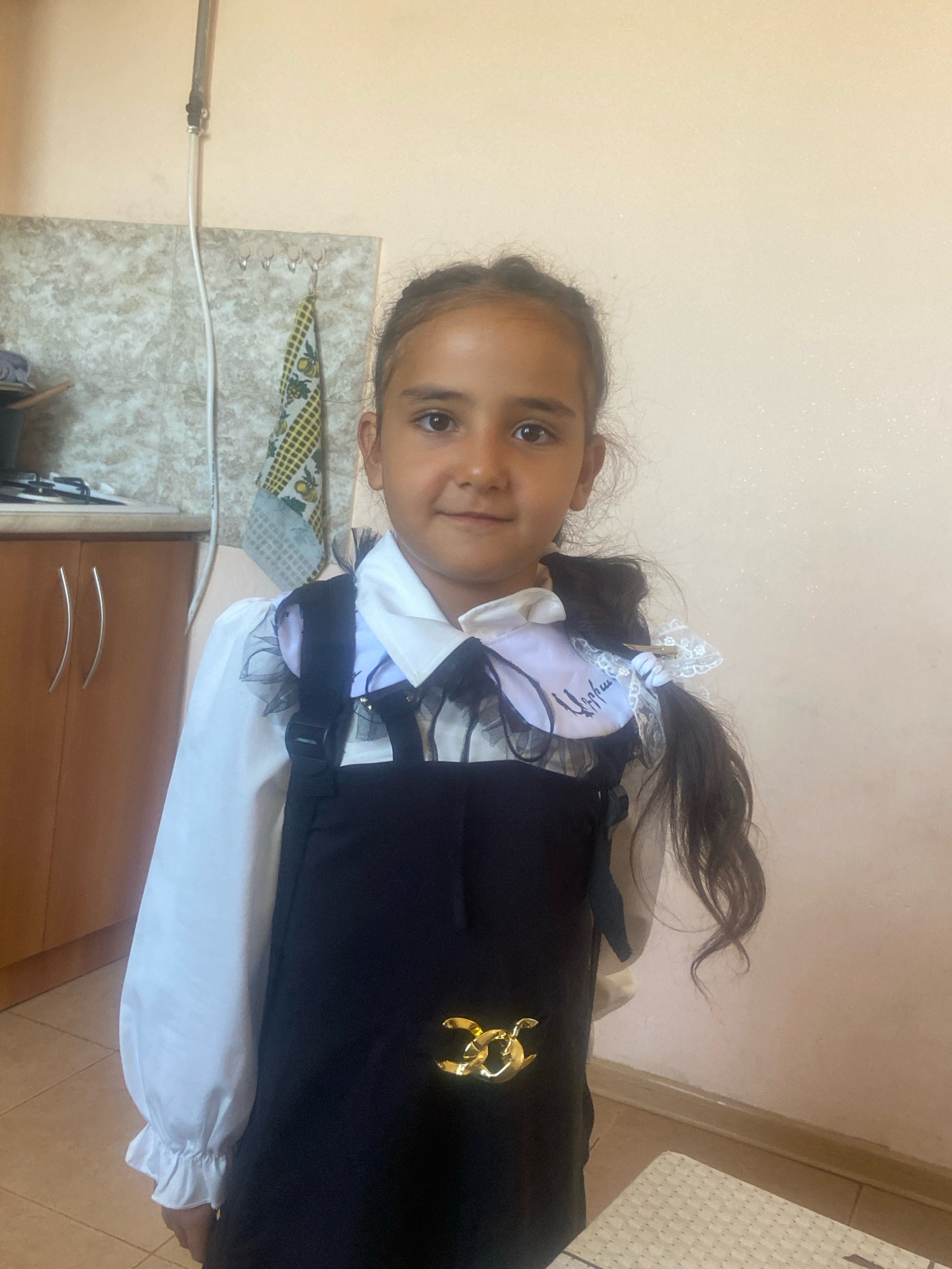 A young girl stands facing the camera in what appears to be a home kitchen. She is wearing a white collared shirt with ruffled sleeves and a black pinafore dress. The dress has a decorative gold chain-like embellishment on the front. Her dark hair is pulled back in a ponytail, adorned with white lace bows, one of which has handwritten text in a script that appears to be Armenian. The background shows a tiled backsplash, kitchen cabinets with modern handles, and a neutral-colored wall. A pipe runs vertically along the wall, and a patterned dish towel hangs from hooks. The lighting suggests it is daytime. The girl has a neutral expression.