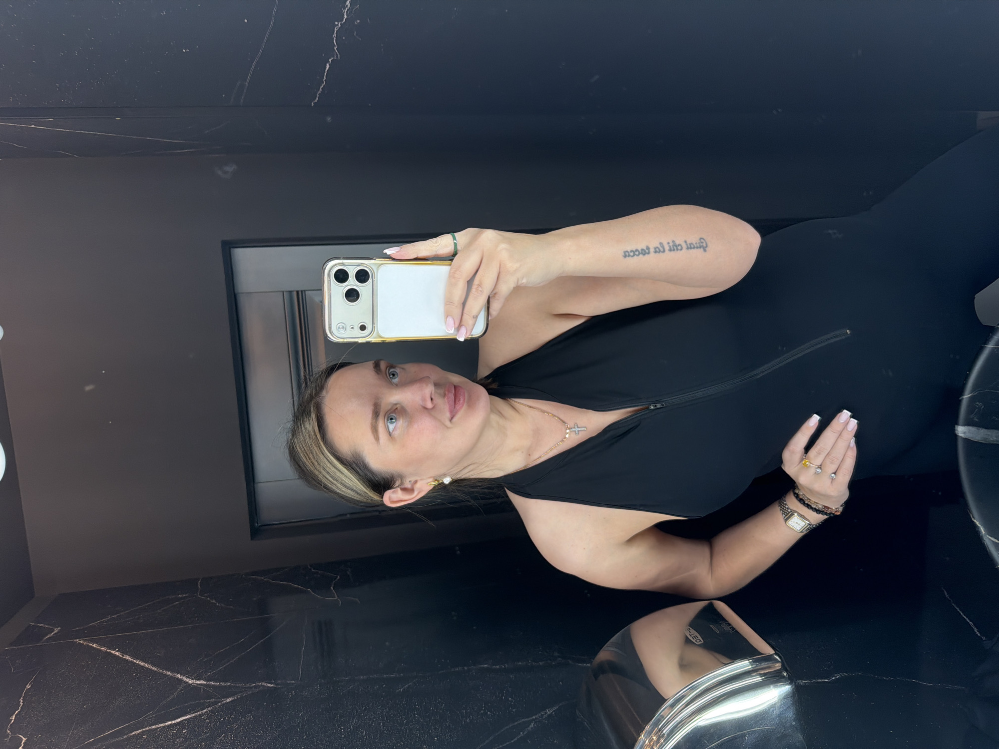 A woman is taking a selfie in a bathroom. She is wearing a black sleeveless top with a zipper. She has a gold necklace with a cross pendant, a watch, and several bracelets on her right wrist. She is holding a phone with a gold case in her left hand. The bathroom has black walls and a black marble countertop. The image provides no information indicating the location in the city of Moscow, Russia.