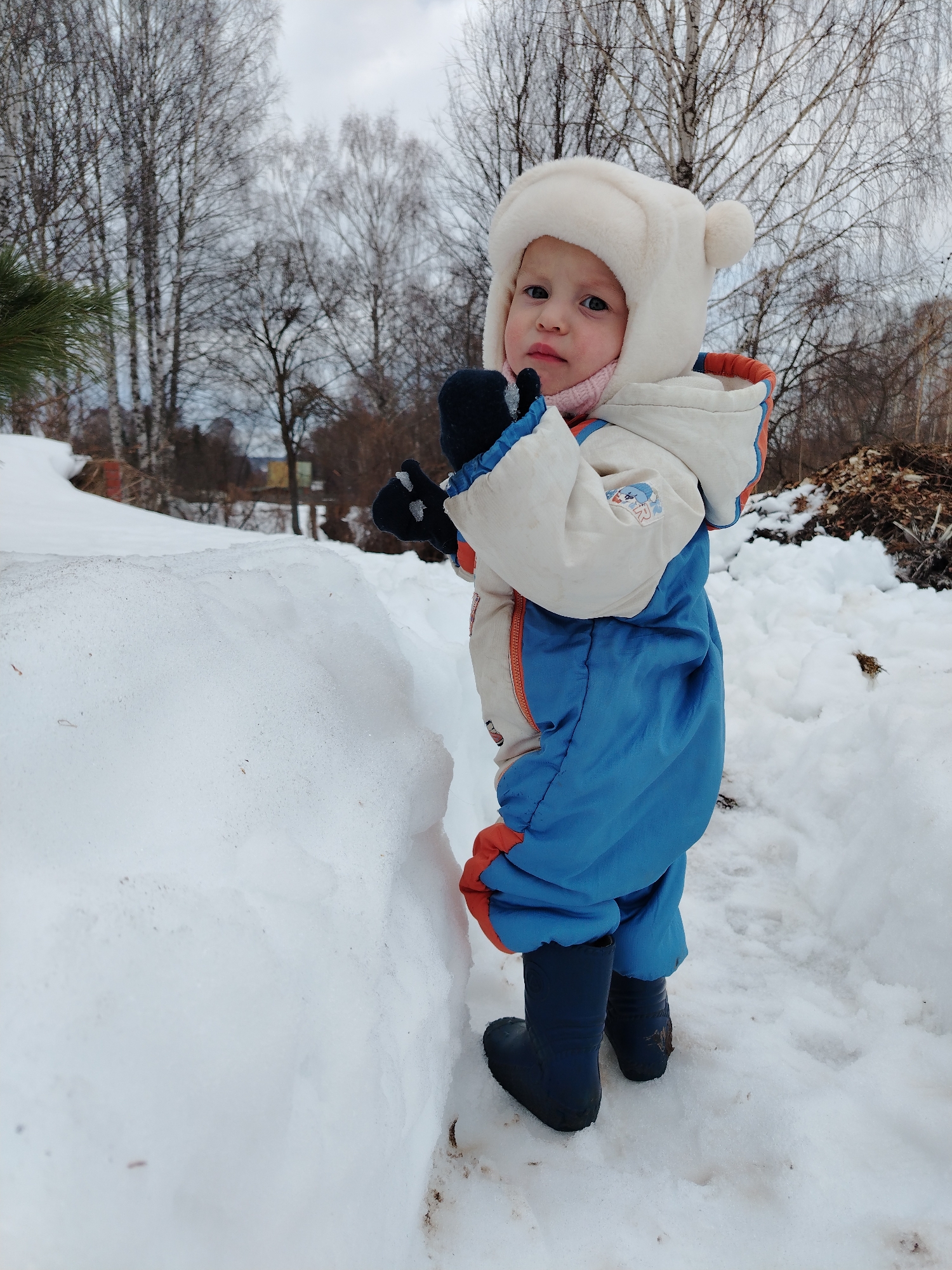 A young child stands in a snowy environment. The child is wearing a white winter hat with pom-poms, a blue and white snowsuit with orange trim, and dark rubber boots. The child is standing next to a large snowdrift, with their hands in black gloves near their face. The background includes bare trees and a pile of brush. The scene is in Nizhnyaya Gar', Russia.