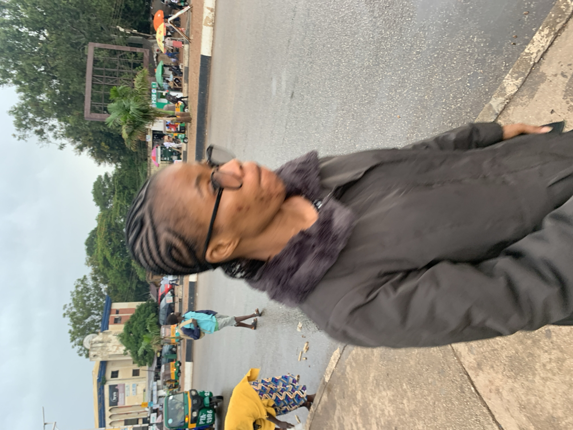 A medium shot shows a young woman with braided hair and glasses walking outdoors on a street, with her body angled towards the right of the frame. She is wearing a dark jacket with a furry collar. In the background, several people, a green and yellow auto-rickshaw, and buildings are visible. The street appears wet, indicating recent rain. The woman is the primary subject, and the environment suggests a bustling urban area.