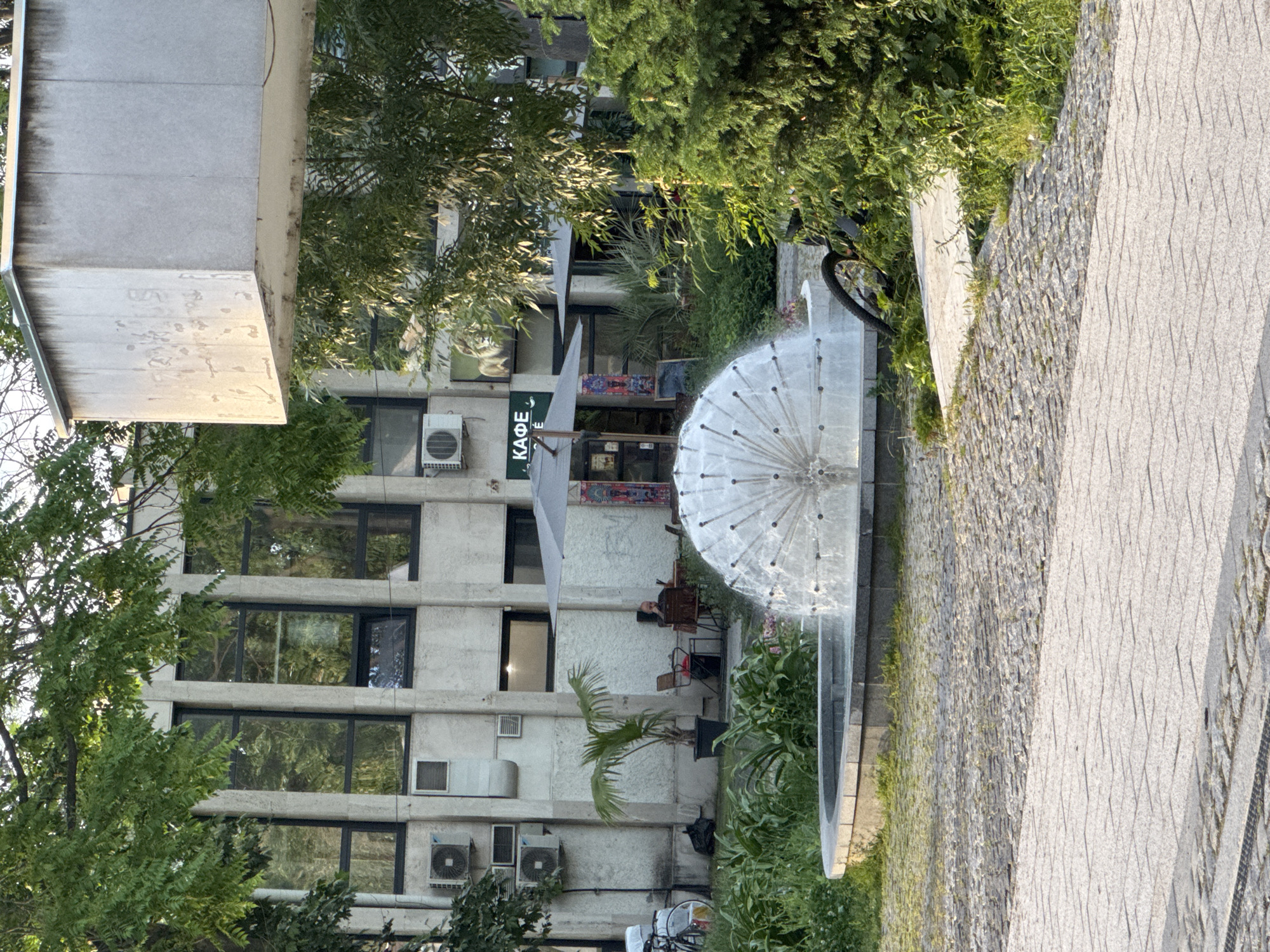 A scene in Plovdiv, Bulgaria, features a multi-story concrete building with numerous windows and air conditioning units attached to its exterior. A fountain with a dandelion-shaped spray sits in front of the building, surrounded by greenery and a paved pathway. A cafe is visible with outdoor seating occupied by people. A bicycle is parked near the fountain.