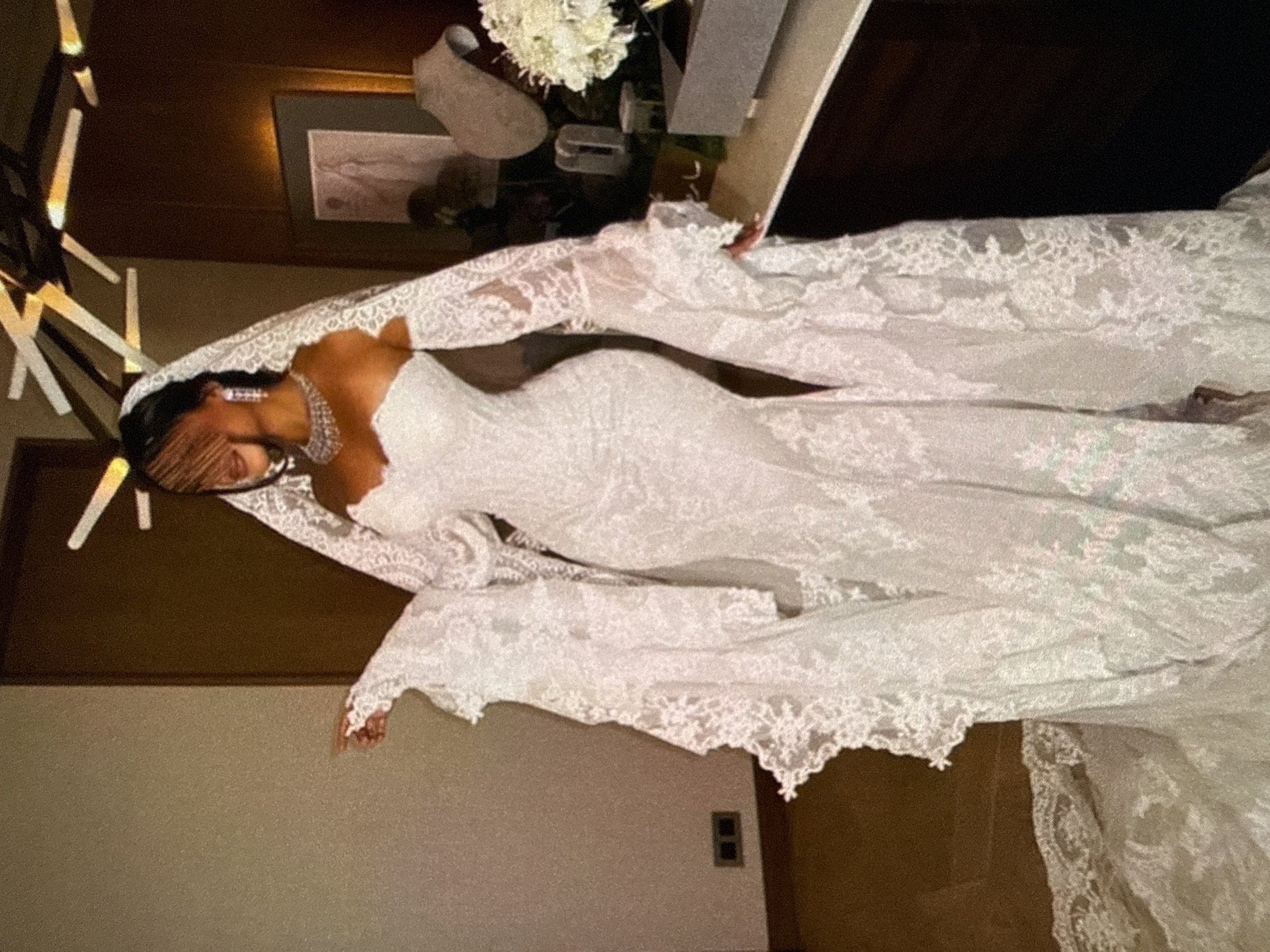 A woman is wearing a white lace wedding dress and veil. She is also wearing a diamond necklace and earrings. The location appears to be indoors. A modern light fixture and framed artwork are partially visible in the background. White flowers are visible on a nearby table.