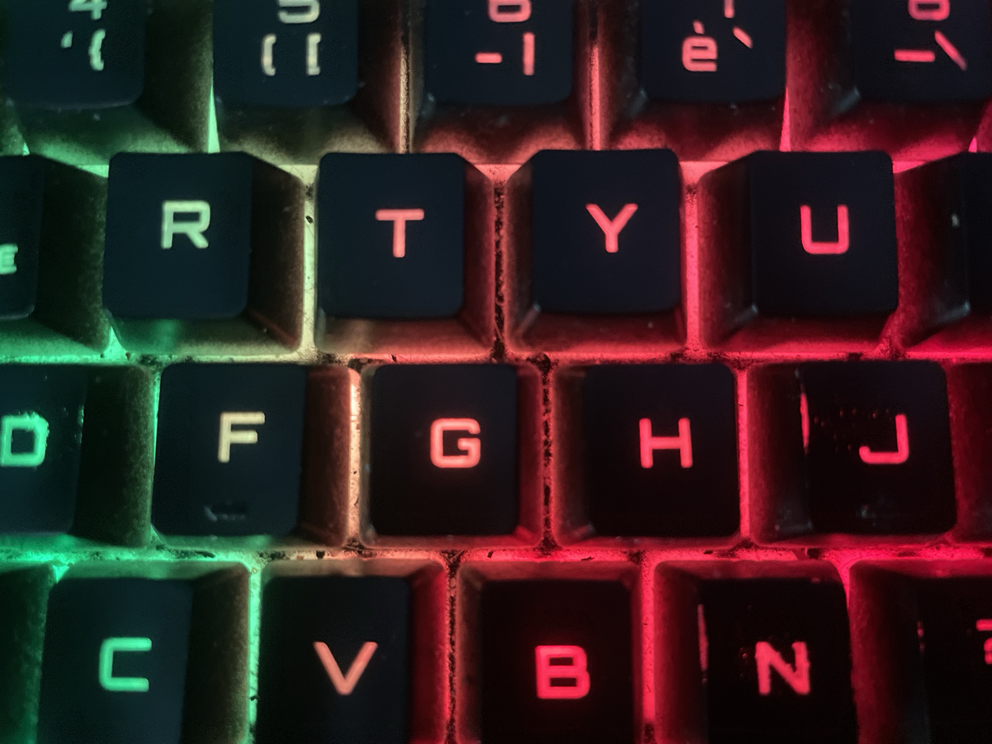 This is a close-up, high-angle, top-down shot of a keyboard, likely a gaming keyboard due to the illuminated keys. The keys are black with white or red lettering. The lighting on the keys shifts from green on the left side of the image to red on the right, suggesting a customizable RGB lighting effect.
The visible letters on the keys, from top to bottom and left to right, include: '4', '}', '{', 'é', '-', '1', 'R', 'T', 'Y', 'U', 'D', 'F', 'G', 'H', 'J', 'C', 'V', 'B', 'N', and what appears to be a stylized symbol resembling 'コ' or 'L'. Some keys have faint smudges or debris. The scene is an indoor setting, focusing solely on the keyboard itself. There is no indication of time of day, weather, or specific location beyond the assumption of a gaming setup. The emotions evoked are neutral, given the lack of human presence. The text visible is solely the characters on the keys.