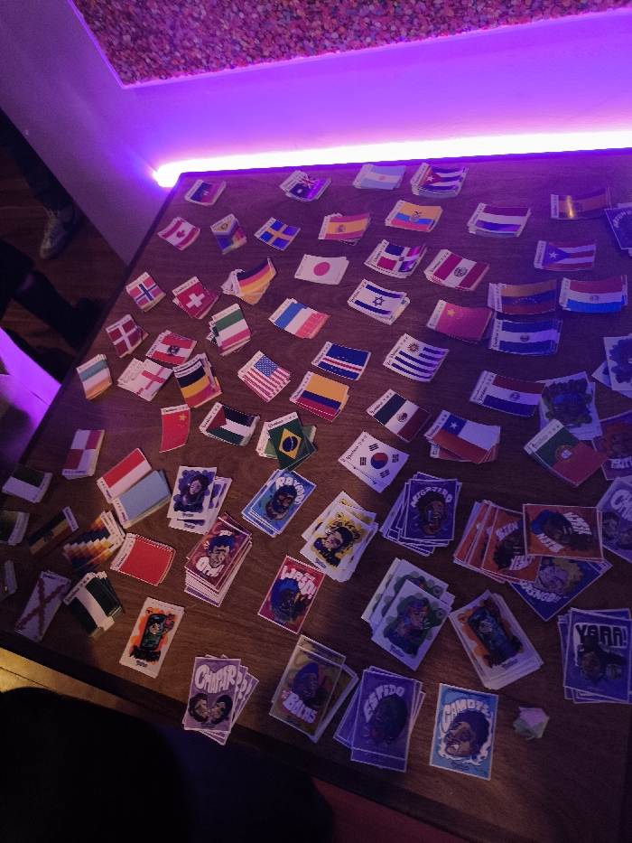 The image depicts an indoor scene in La Paz, Bolivia, featuring a light-brown wooden tabletop illuminated by purple LED strip lighting mounted on an adjacent wall. Above the LED strip, a textured ceiling or wall segment is visible, adorned with numerous small, brightly colored, pebble-like or confetti-like elements.
The tabletop is densely covered with hundreds of small, rectangular paper stickers or cards, predominantly arranged in a scattered manner, with some in small stacks. A significant portion of these items are national flags, including those of the USA, Canada, Japan, Sweden, Germany, Israel, Mexico, Chile, Brazil, South Korea, Argentina, Spain, Norway, and the United Kingdom. Interspersed among the flags are numerous illustrated cards featuring stylized cartoon characters, accompanied by bold, capitalized Spanish text such as "RAYADO," "NEGATERO," "WARAO!," "CHUPAR," "CAMOYO," "ESVIDO," "FIESTERO," "BINGOS," "YARRA!," and "LAS BOLAS." A small, irregularly shaped, light-green object rests on the table in the lower right quadrant.
In the upper left background, the lower legs and feet of an individual wearing dark pants and gray athletic shoes are partially visible. A dark, indistinct form, consistent with a person's leg or body, is barely visible in the lower left foreground. No specific actions or interactions between individuals are discernible. The precise venue within La Paz, Bolivia, is not identifiable from the image content.