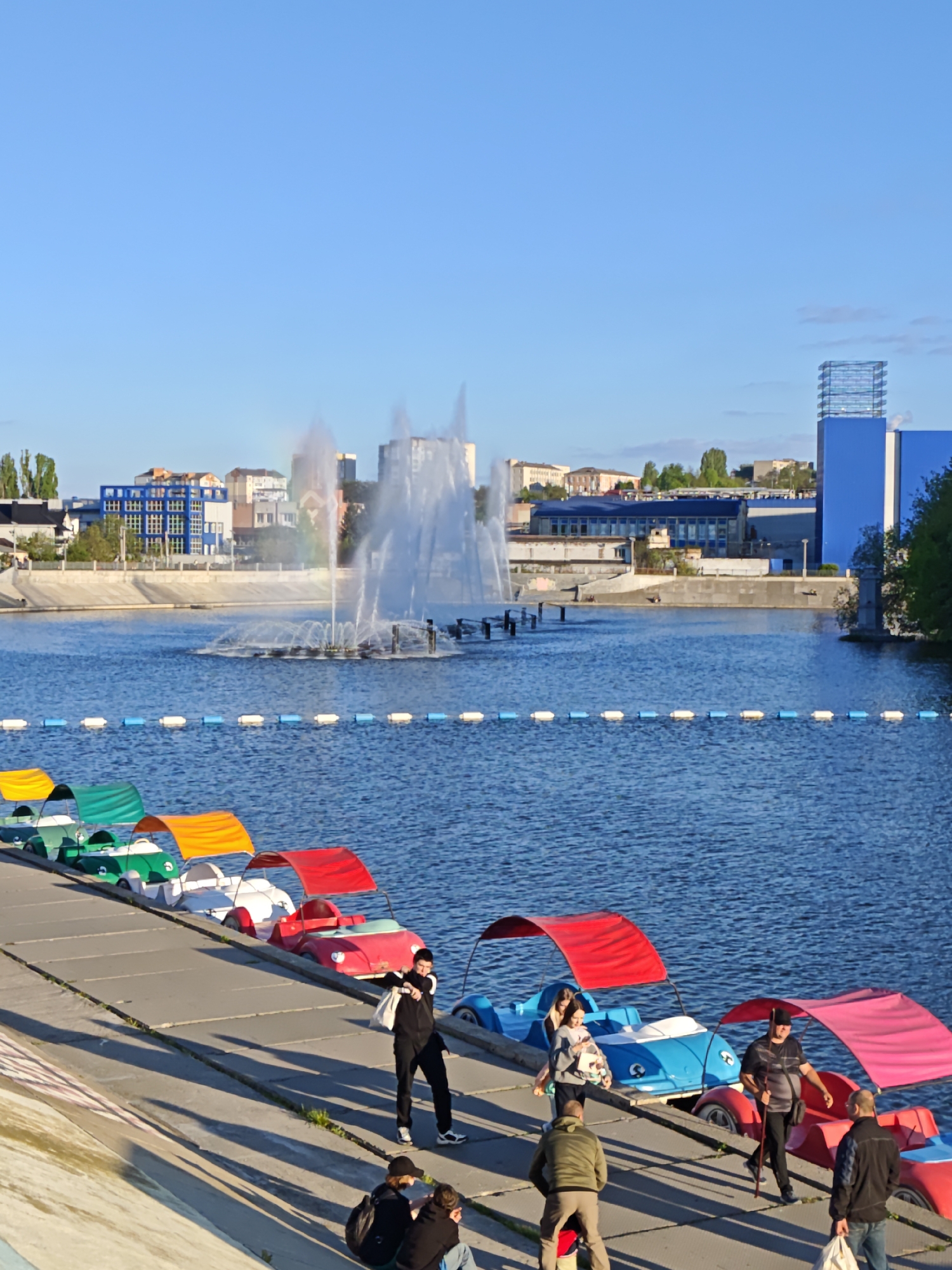 The image shows a waterfront scene in Vinnytsia, Ukraine. In the foreground, there is a line of colorful paddle boats shaped like vintage cars. People are gathered on the concrete embankment where the boats are docked. Some are standing, while others are sitting on the embankment, near the boats.
In the background, across the water, there are buildings including one with a blue facade and exposed metal framework. A fountain in the middle of the water shoots multiple streams high into the air. A partial rainbow is visible within the water spray of the fountain. The sky is clear and blue.