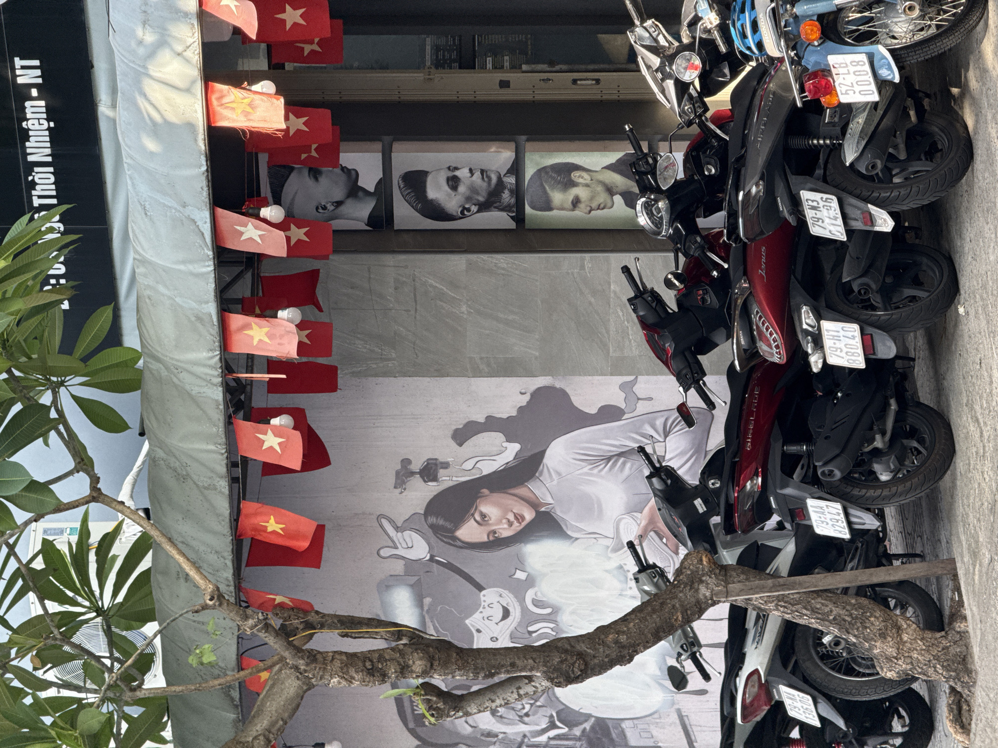 In a street scene in Nha Trang, Vietnam, a row of parked scooters is visible in the foreground. Behind the scooters, a mural depicts a woman in traditional Vietnamese attire holding a white object, possibly a piece of clothing. The mural is adorned with small red Vietnamese flags. To the right of the mural, three framed portraits of men with stylized hairstyles are displayed, suggesting a barbershop. The building above features signage with Vietnamese text. The overall environment appears to be an urban commercial area, with a tree contributing to the foreground elements of the composition.