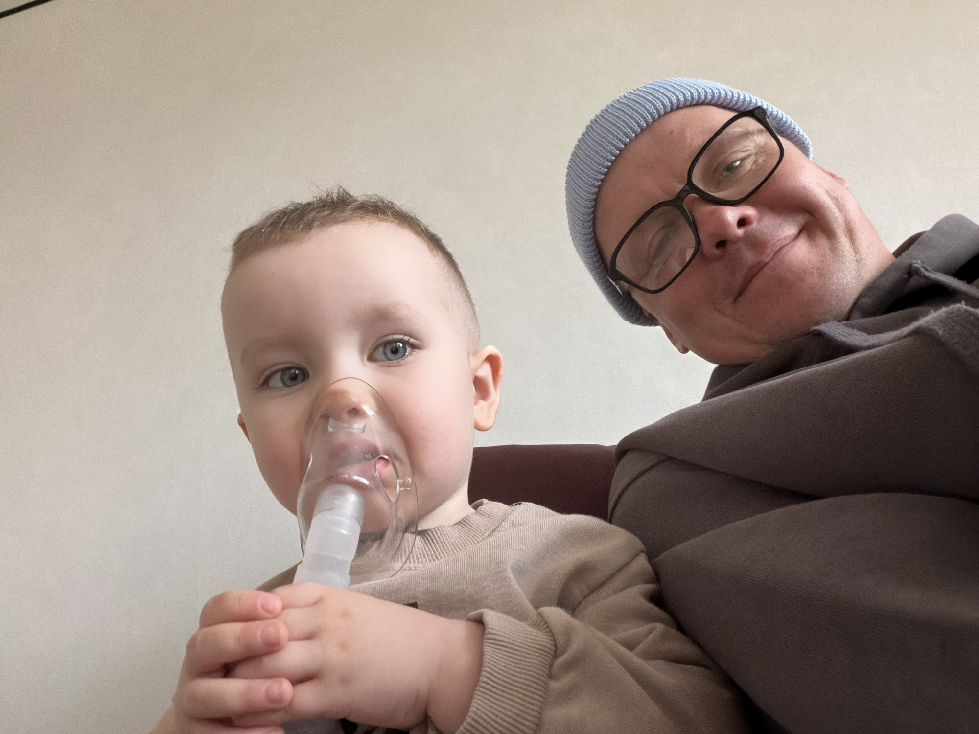 A close-up shot reveals a toddler, likely around 2-3 years old, wearing a clear plastic nebulizer mask over his nose and mouth. He holds the base of the nebulizer in his hands. He has short, light-colored hair and blue eyes. Adjacent to the child is an adult male, presumably a father or guardian, wearing glasses and a light blue knit cap. The adult is positioned slightly behind and above the child, partially obscured. Both are wearing casual clothing. The background is a plain, light-colored wall. There are no indications of the location in the city of Chaikovsky, Russia.