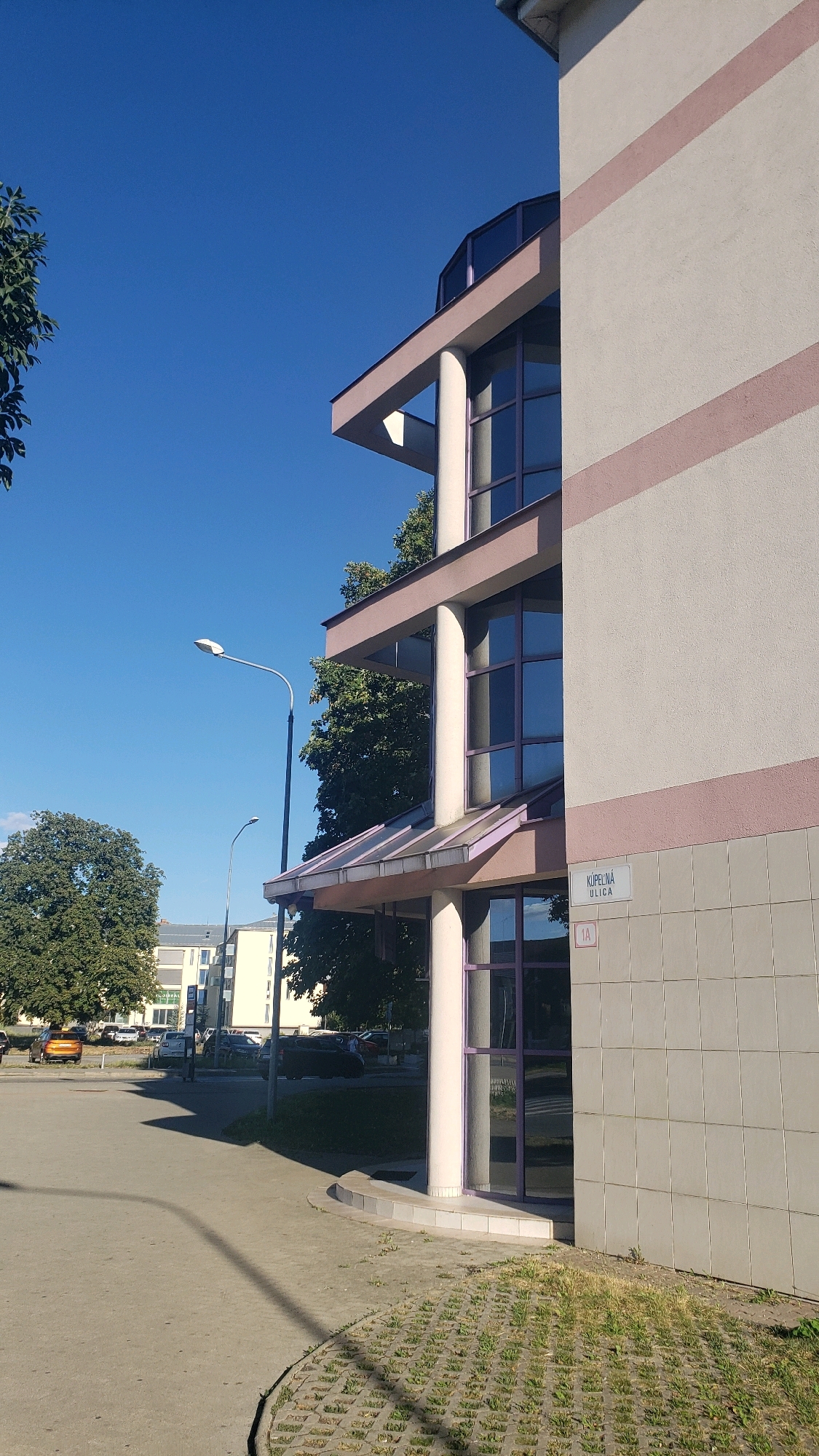 The image shows the exterior of a modern building in Prešov, Slovakia. The building has a light-colored facade with horizontal pink stripes. It features multiple levels with large, reflective windows framed in purple. A street sign on the building reads "Kúpeľná ulica 1A". A large tree is visible to the left of the building, and in the background, other buildings and parked cars can be seen. The sky is clear and blue, and a street lamp stands on the left side of the image. The ground is a combination of paved areas and permeable paving with grass. The scene appears to be a public space, possibly a commercial or residential area.